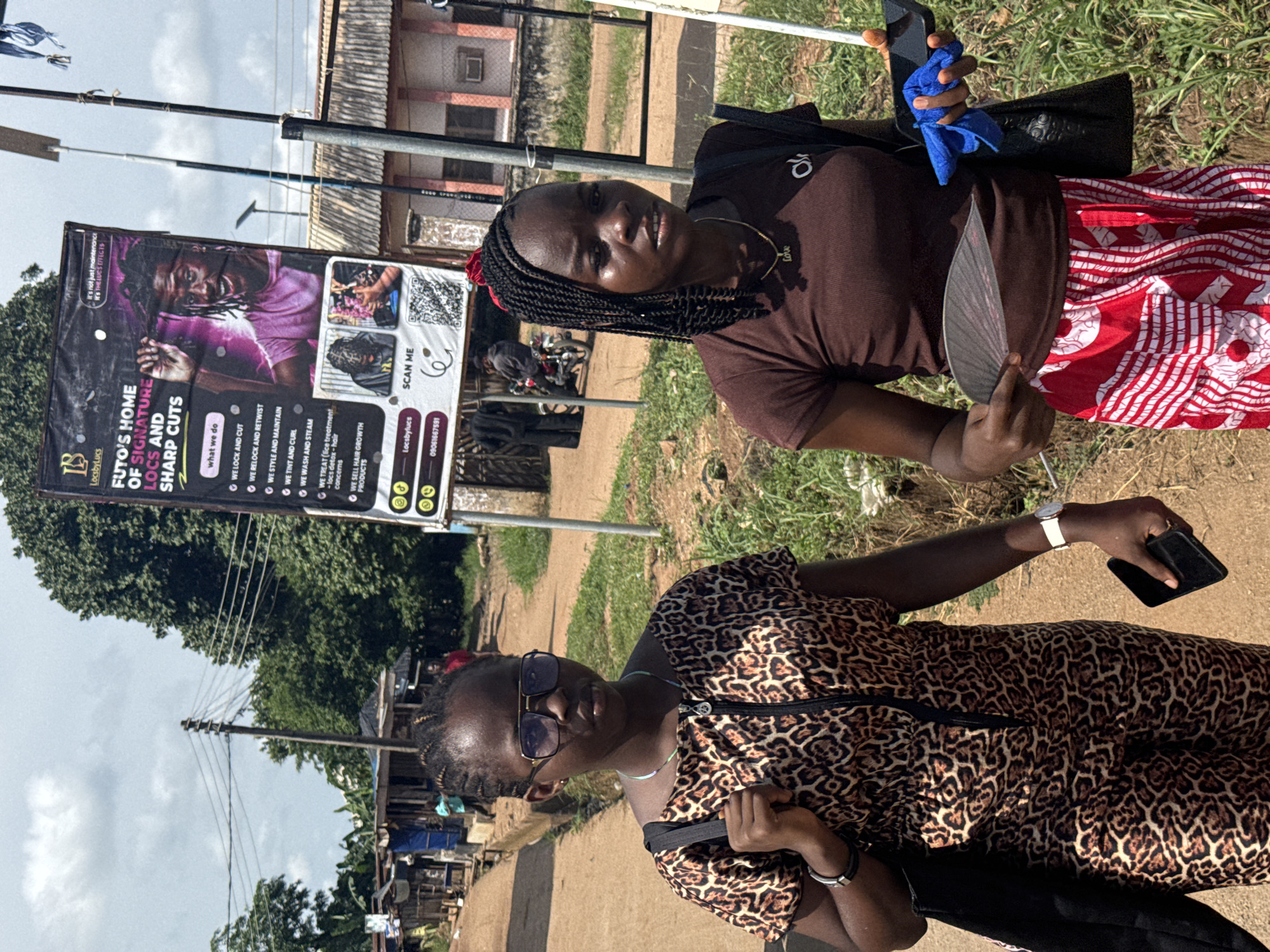 Two young women are standing outdoors on what appears to be a dirt road or path, with a large business banner behind them, suggesting a street scene in Ihiagwa, Nigeria. The sky is partially cloudy, and the overall setting includes sparse foliage, utility poles with wires, and simple buildings in the distance.
The woman on the left wears a leopard-print short-sleeved dress, dark sunglasses, and has braided hair. She holds a black smartphone in her right hand and a black bag is visible on her left shoulder. A white watch is on her left wrist, and a bracelet on her right.
The woman on the right wears a brown short-sleeved t-shirt and a red and white patterned skirt. She has braided hair, with some red accents, and wears a necklace with the word "Love" as a pendant. She is holding a dark-colored fan in her right hand and a black smartphone, wrapped in a blue cloth, in her left hand. Both women are looking towards the camera with neutral to slightly smiling expressions.
A prominent banner in the background advertises "FUTO'S HOME OF SIGNATURE LOCS AND SHARP CUTS" by "LocsbyLucs." The banner lists services such as "WE LOCK AND CUT," "WE RELOCK AND RETWIST," "WE STYLE AND MAINTAIN," "WE TINT AND CURL," "WE WASH AND STEAM," "WE TREAT (locs treatment - locs detox - hair concerns)," and "WE SELL HAIR GROWTH PRODUCTS." It also features the slogan "it's not just maintenance IT'S THE LOCS EFFECTS," a QR code for "SCAN ME," and a contact number "09061667591." The presence of the fan suggests warm weather.