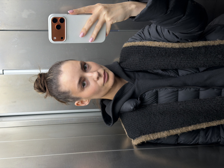 A young adult female is depicted in a medium shot, taking a selfie inside an elevator in Unknown, Unknown. She has dark brown hair pulled into a messy bun and a neutral facial expression, looking directly at the camera. She is wearing a black hooded sweatshirt under a black puffer jacket, with a black knitted scarf featuring a prominent light brown stripe draped over her shoulders. Her left hand holds a light-colored smartphone, possibly white or light grey, encased in a protective cover with an orange-brown rectangular module housing three camera lenses. The phone case displays a "TRON" logo. Her fingernails are manicured with light pink polish. The elevator interior features vertical brushed metal panels.