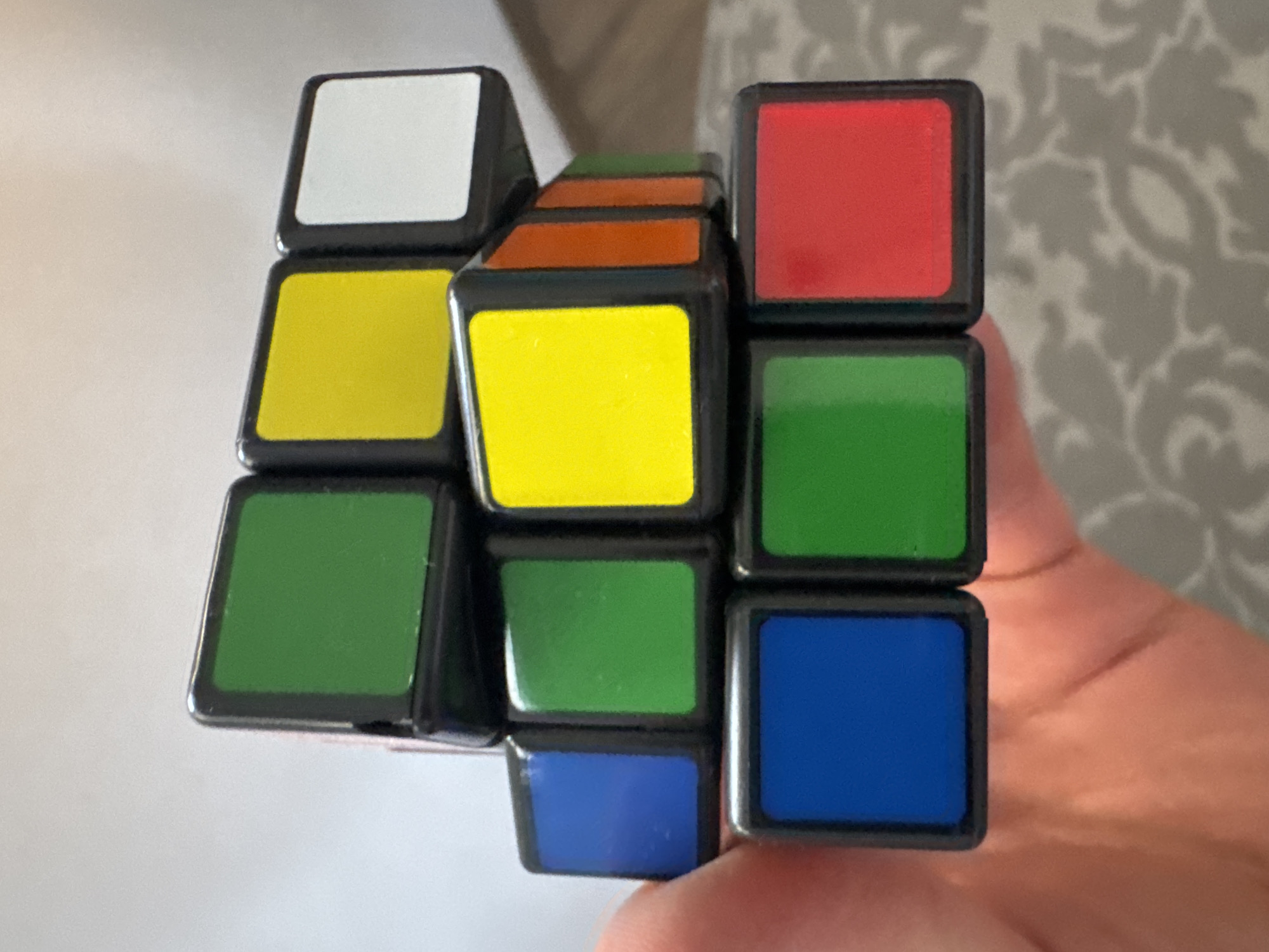 A Rubik's cube is held by a person's hand against a light-colored background and a patterned wallpaper. The cube is partially solved, with some faces showing their standard colors and others in mixed states. The visible colors on the cube are white, yellow, green, red, and blue, arranged in a 3x3 grid. The image is taken from a slightly elevated angle, looking down at the cube. The lighting suggests it is daytime, likely indoors, as there are no strong shadows indicating direct sunlight. The setting is domestic, given the wallpaper and the presence of a hand holding a common toy. The overall mood is neutral, focusing on the object itself. There is no visible text in the image.