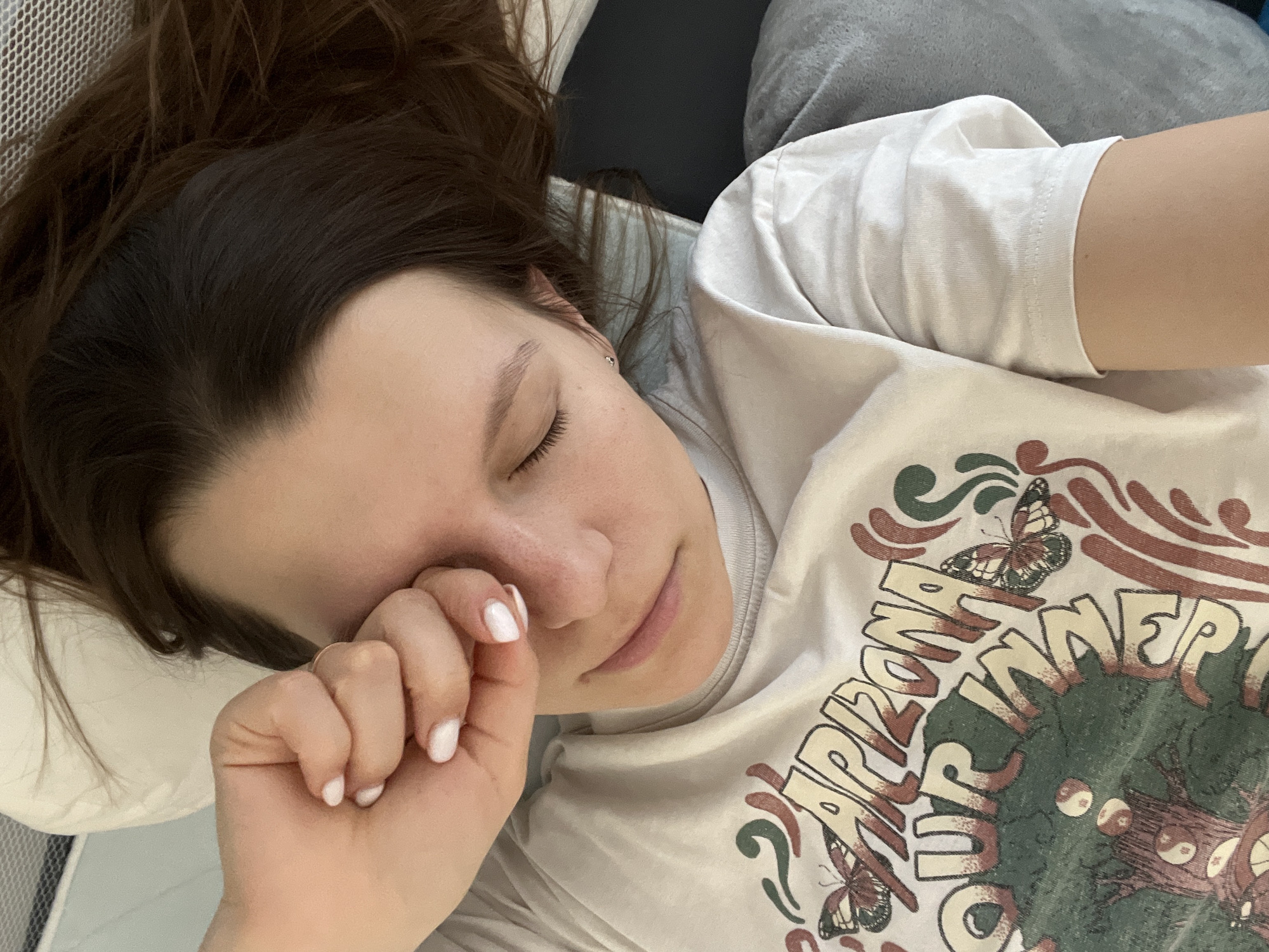 A close-up, overhead shot shows a young person with their eyes closed, resting their head on a pillow. Their hand is raised to their face, with their fingers lightly touching their cheek. They are wearing a white t-shirt with a graphic design featuring text and imagery of a tree and butterflies. The text on the shirt reads "ARIZONA OUR INNER". The scene appears to be indoors, likely in a bedroom or a casual setting, with hints of a bed or couch in the background. The lighting suggests it could be daytime, with soft illumination. The person's expression and posture convey a sense of relaxation or sleepiness. The overall mood is peaceful and intimate. The visible text on the shirt suggests a possible connection to Arizona or a brand with that theme. The location context of Limassol, Cyprus, is provided but not directly evident in the image itself.
