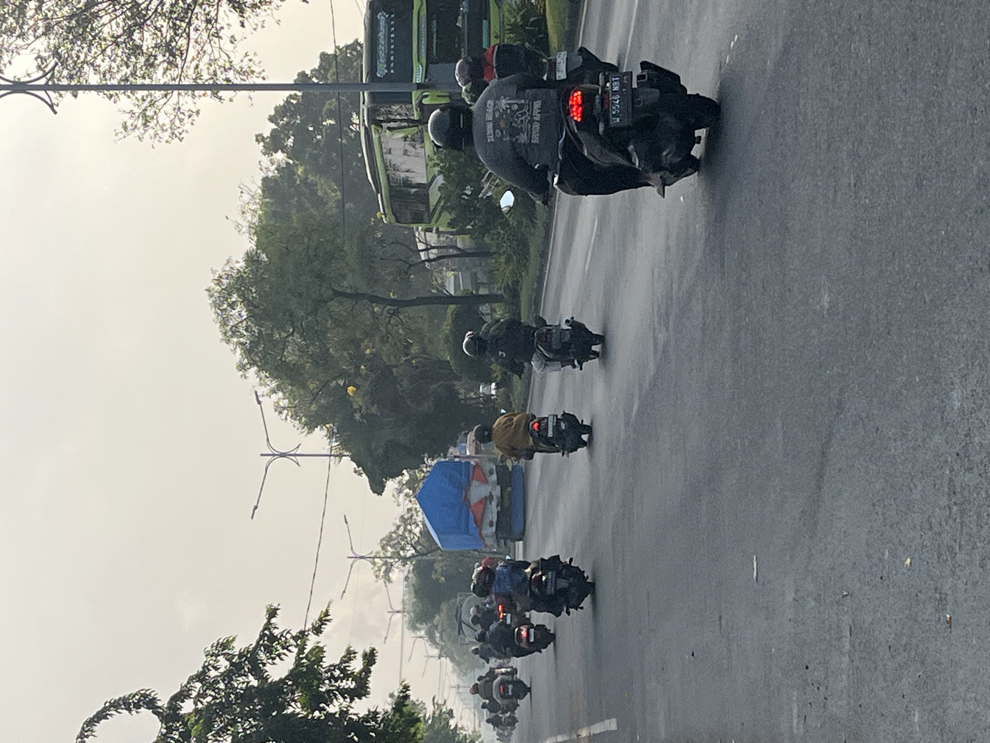 The image depicts a street scene in Gelam, Indonesia, with a procession of motorcycles on a paved road. The sky is overcast and hazy, suggesting it might be early morning or late afternoon. Several motorcycles are in motion, some with riders wearing helmets. In the background, a large truck covered with a blue tarp is visible, along with trees and what appears to be a green bus. Lampposts are also present, lining the street. The overall atmosphere is that of everyday traffic in an urban environment.