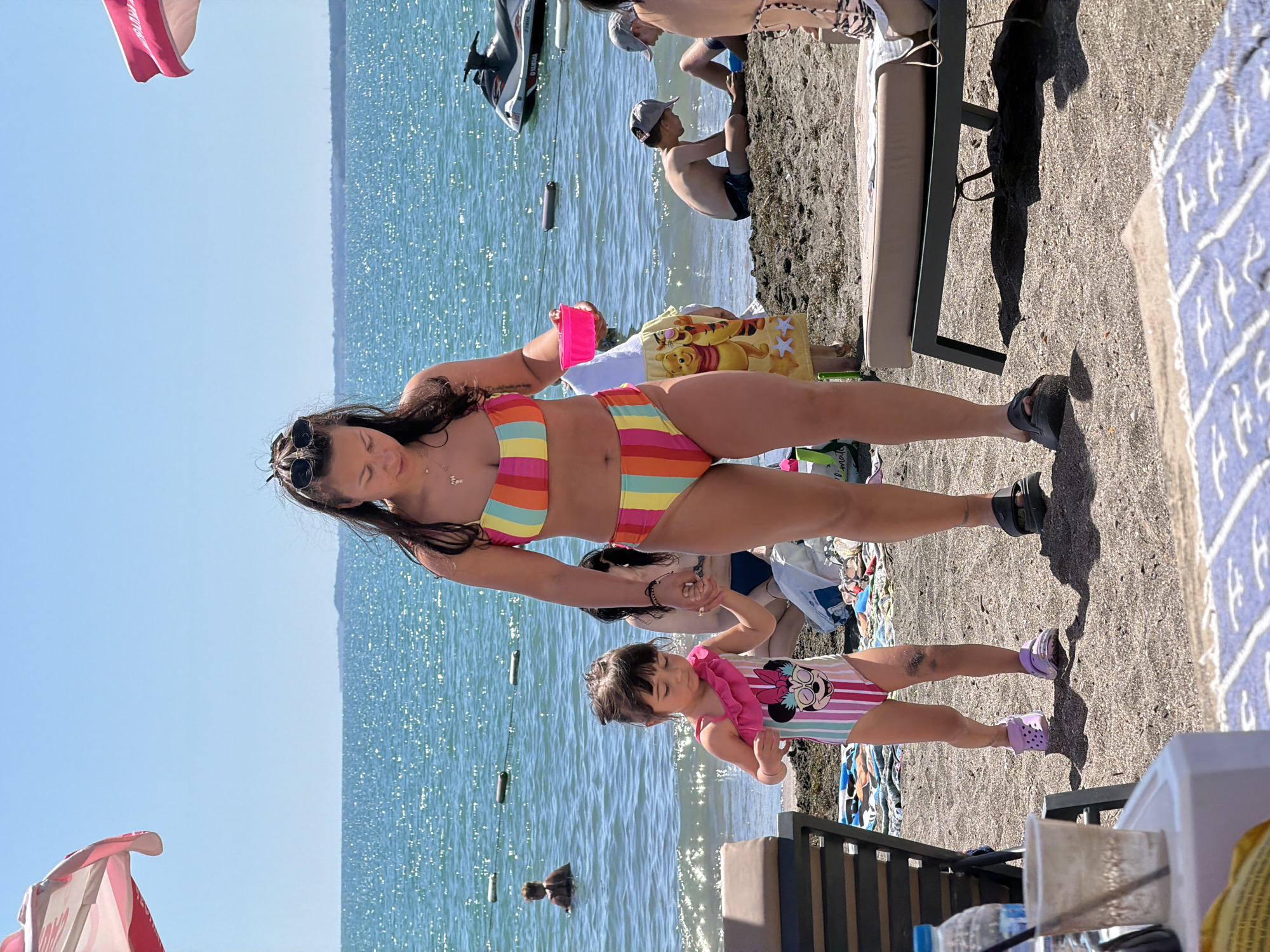 This media file depicts a sunny day at a beach in Burgas, Bulgaria. The main subjects are a woman and a young girl, both wearing swimwear. The woman is in a colorful striped bikini, and the girl is in a swimsuit with a Minnie Mouse design. They appear to be engaged in a playful interaction, with the woman holding the girl's hand.
The scene is set on a sandy beach adjacent to a body of water, likely the Black Sea, given the location context. The water is calm and reflects the bright sunlight, creating a shimmering effect. In the background, several other people are visible, some in the water and others on the beach. A jet ski is parked on the shore, suggesting recreational activities are available. Beach chairs and towels are scattered around, indicating a typical beach day.
The overall mood is relaxed and cheerful, consistent with a vacation setting. The bright sunlight and clear blue sky suggest fair weather, likely during the daytime.
Visible text includes "YAMAHA" on the jet ski and "MINNIE" on the girl's swimsuit. There is also some text on a pink and white towel in the upper left corner of the image.