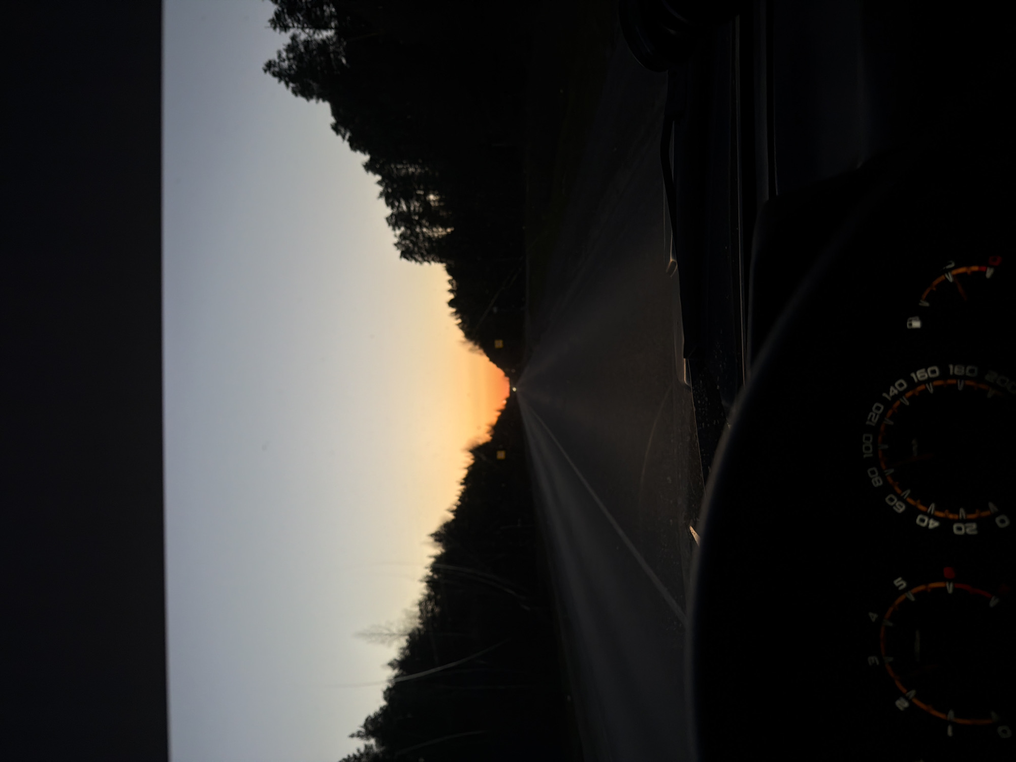 The image shows a view from inside a car driving on a paved road, likely in Chaikovsky, Russia. The sun is setting in the distance, casting an orange glow on the horizon. Trees line both sides of the road. The car's dashboard is visible, including the speedometer and fuel gauge. The speedometer needle points towards 0 km/h, suggesting the car may be stationary or moving slowly. The fuel gauge indicates the tank is close to being empty.