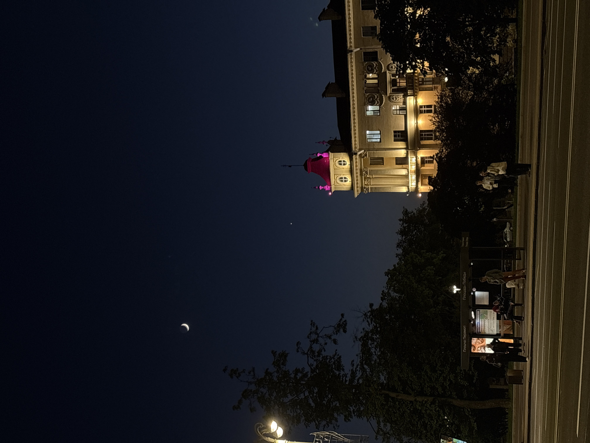 The image shows a night scene in Minsk, Belarus. A building with illuminated windows and a tower topped with pink lighting stands prominently. A crescent moon is visible in the dark sky. Streetlights illuminate the foreground where people are gathered at a bus stop. Trees line the street, and the city's night sky is a deep blue.
