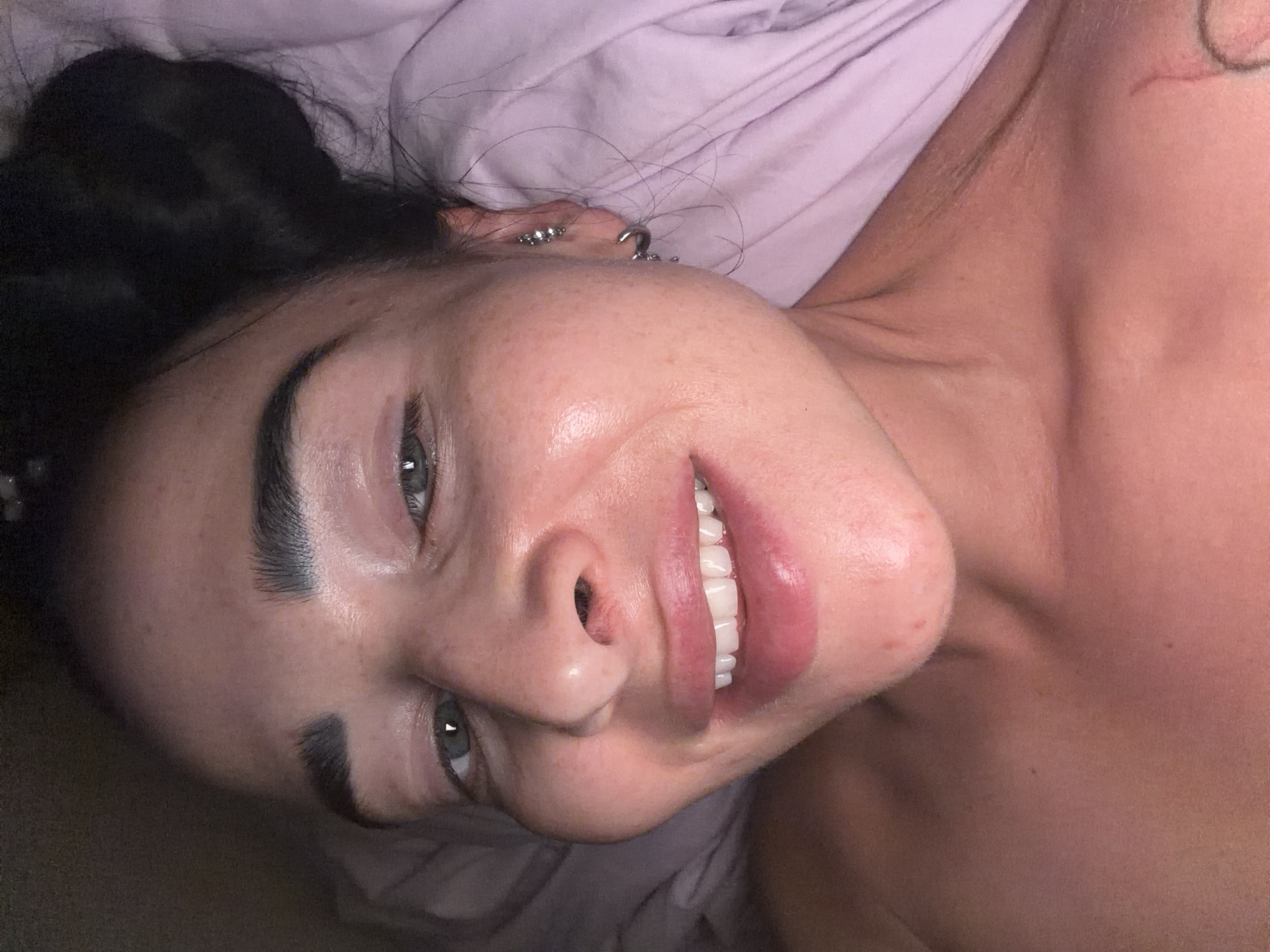 This close-up portrait captures a young woman lying down, looking up and smiling directly at the viewer. Her head is angled, with dark hair, possibly tied up in a bun, visible against a soft, light purple fabric background, likely a bedsheet or pillow. She has dark, well-defined eyebrows and light-colored eyes, possibly blue or grey, which convey a friendly and relaxed expression.
A broad, genuine smile reveals perfectly white, aligned teeth and full lips. Her skin appears clear, smooth, and has a noticeable dewy or glowing finish, particularly on her cheeks and forehead. She has multiple piercings on her left ear, including a conch/helix ring and smaller studs further up the helix. A partial view of a circular tattoo can be seen on her right shoulder/neck area. Her shoulder and upper chest are visible, suggesting she is shirtless or wearing something that exposes her décolletage.
The overall impression is one of contentment and warmth. The lighting is soft and even, highlighting her features without harsh shadows, suggesting an indoor setting. No specific location cues for Saint Petersburg, Russia, are visible within the image itself. There is no visible text.