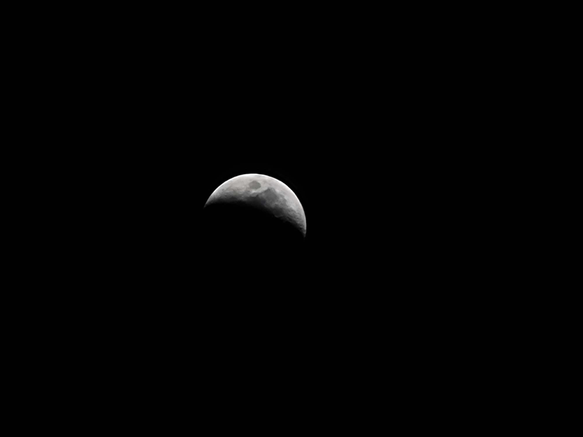 A crescent moon is visible in a black sky. The moon is partially illuminated, with the illuminated portion being a bright white. The unilluminated portion is a deep black, blending into the night sky. The surface of the moon shows some craters and texture. The image is a close-up of the moon, with no other objects or features visible. The location of Quarteira, Portugal, is not depicted in this image.