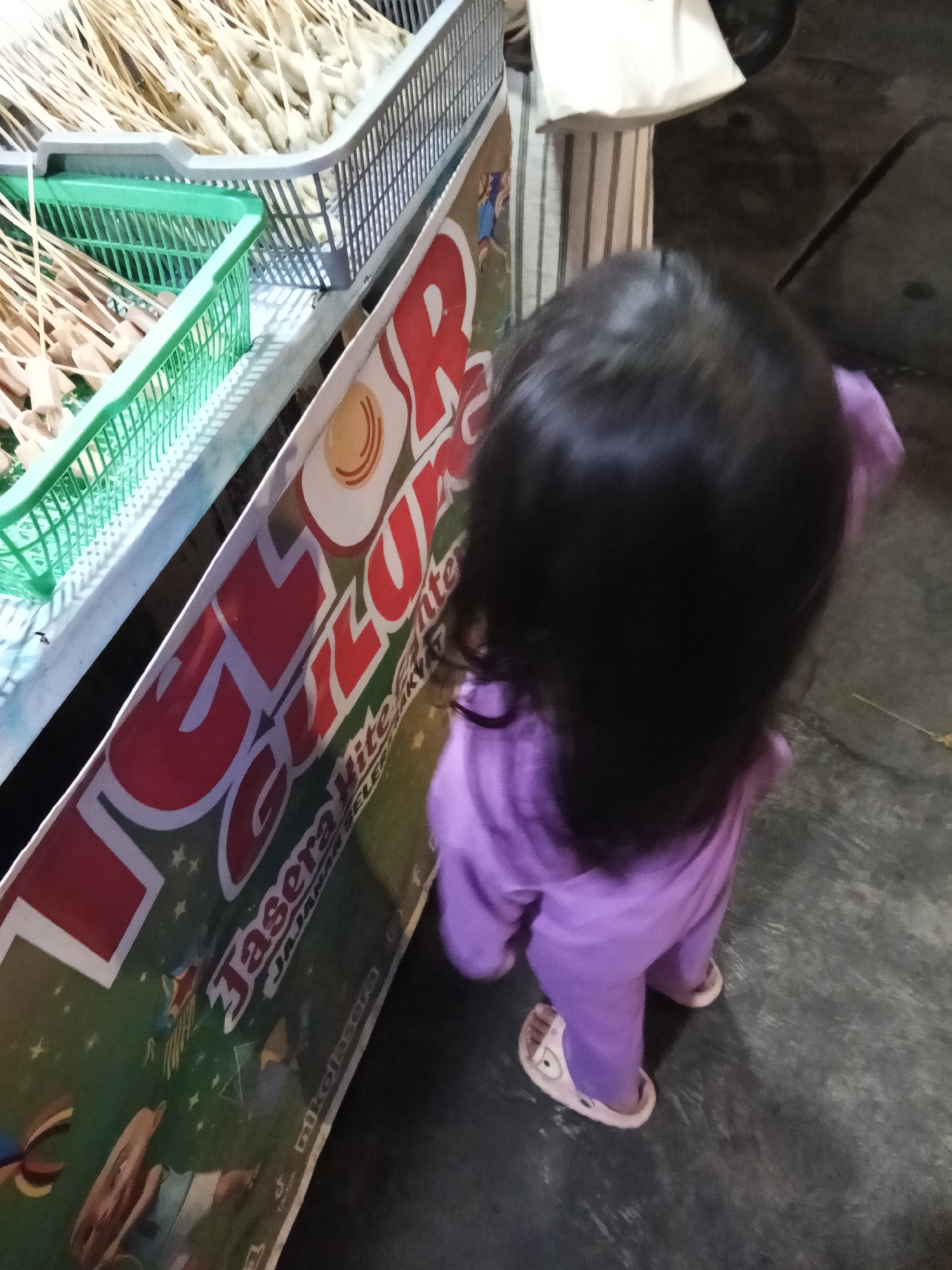 A young child wearing a purple outfit and pink slippers stands on a dark gray paved surface, facing away from the camera towards a food stall. The child's back and head are visible. To the left, a banner with red and white lettering, partially obscured by the child, advertises "JASERA KITE" and "JAJANAN SELE". The banner is green with white stars and images of food and cartoon characters. In the background, various food items on skewers are displayed in green and gray plastic baskets. The overall scene suggests an outdoor market or street food vendor in Lubuklinggau, Indonesia.
