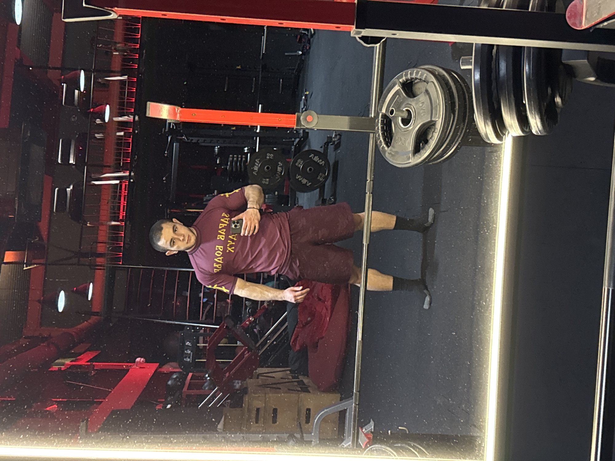 A man stands in a gym, reflected in a mirror. He's wearing a maroon t-shirt and shorts with black socks. He is holding a smartphone in his left hand, presumably taking a photo or video. Behind him is a weightlifting rack loaded with weights. The gym has black floors and red gym equipment. The location is likely Burgas, Bulgaria, based on the Cyrillic script on the t-shirt, though specific city landmarks are not visible to confirm.