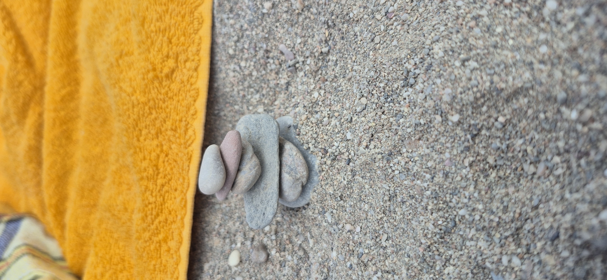 A close-up, eye-level shot shows a stack of five smooth, rounded pebbles balanced precariously on a sandy beach in Paleo Faliro, Greece. The pebbles range in color from light gray to pinkish-gray. Behind the pebbles, a vibrant yellow terrycloth towel is partially visible, suggesting a beach setting. The background consists of granular sand and smaller pebbles, creating a textured surface. The lighting appears to be natural daylight, with a slight overhead angle.