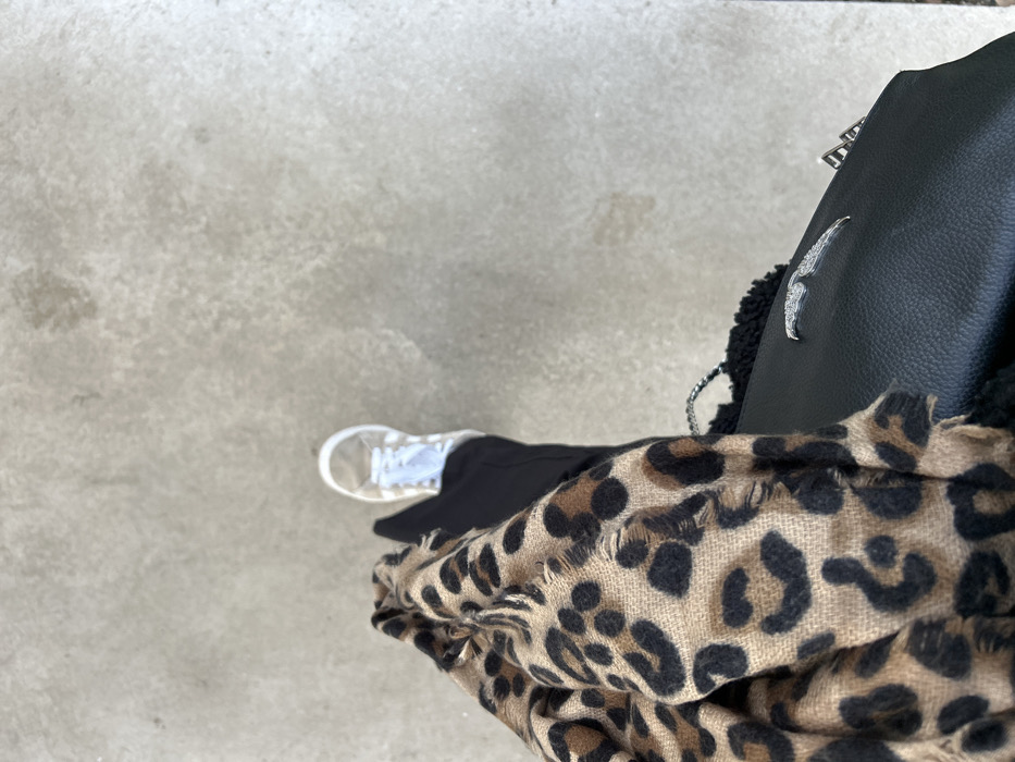 The image displays an overhead, close-up perspective of a partially visible individual standing on a light grey, textured ground surface in Saint-Étienne, France. The lower right leg of the individual is dressed in black trousers, with the right foot encased in a white sneaker featuring white laces. A tan and black leopard print scarf, exhibiting frayed edges, is draped across the lower left quadrant of the frame. Adjacent to the scarf and below the visible leg, a black textured handbag is present, adorned with a distinct silver-toned winged emblem on its exterior. No other subjects or specific environmental identifiers are visible within the frame.