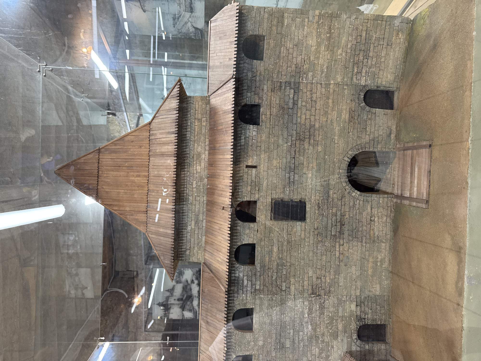 This is a close-up, slightly angled photograph of a scale model of a fortress or castle, likely displayed behind glass. The model is constructed primarily from wood and what appears to be textured material resembling stone or brick for the walls. The main subject is a multi-tiered structure with a prominent wooden, pyramidal roof on the left side. This roof is made of planks and has a serrated edge. Adjacent to the wooden roof is a taller, block-like structure with a brick-like texture. This section has several arched openings or windows, some of which are dark and appear to be doorways or deep recesses. There is also a small rectangular opening on an upper level. A wooden walkway or ramp leads up to an arched entrance on the right side of the brick structure. The ground surrounding the model is a sandy, earth-like texture.
The lighting in the scene comes from above, likely from artificial sources, as evidenced by the reflections on the glass and the bright, elongated white streaks across the top of the image. The background, visible through the glass, is out of focus and appears to be a museum or exhibition space, with hints of other displays or exhibits. The overall impression is that of an architectural model of a historical building, possibly a defensive structure. There is no visible text in the image. The photograph is taken in a way that suggests the model is part of a larger display or exhibit within a building.