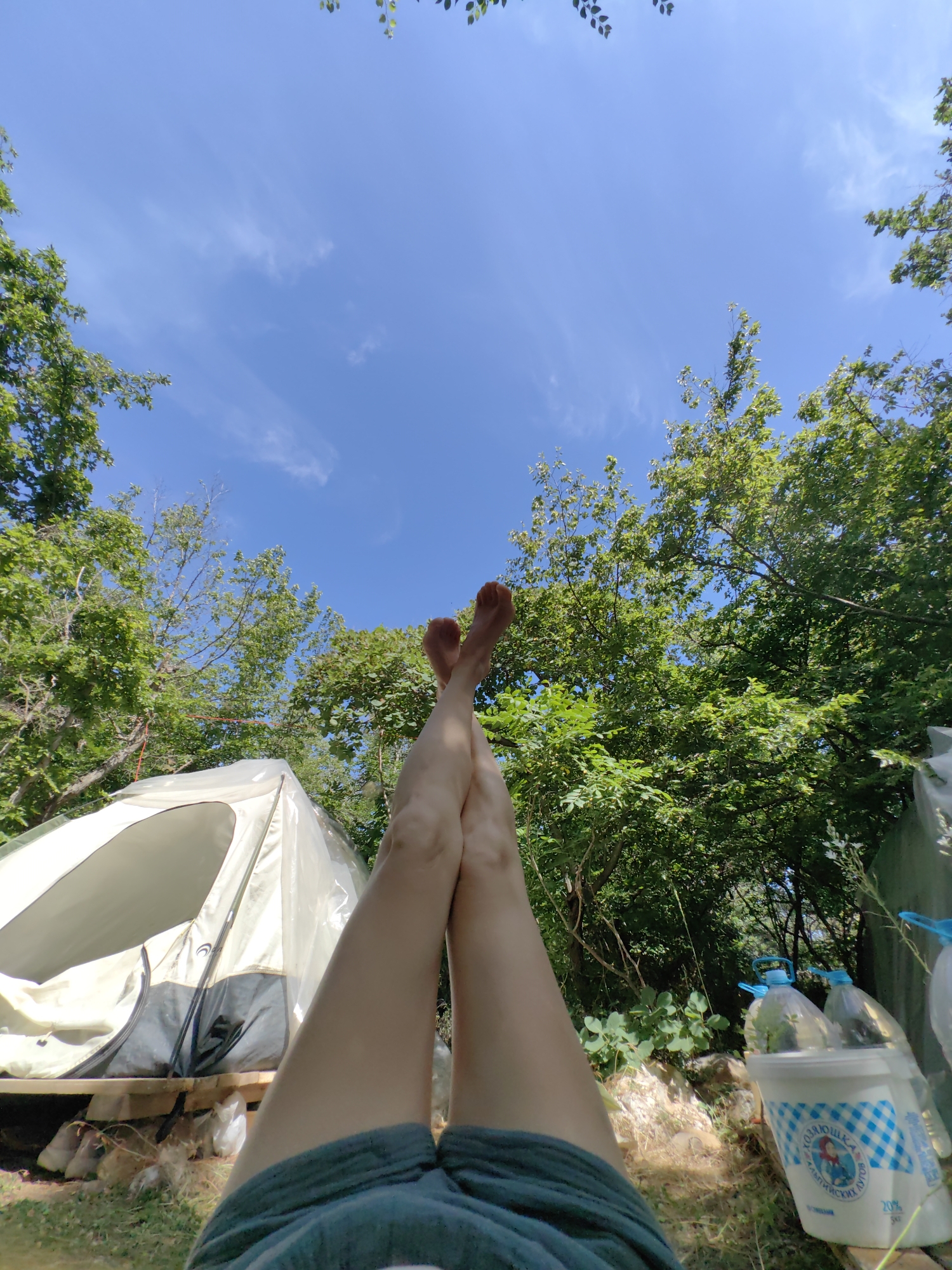 In the city of Utios, Ukraine, a person is lying down outdoors, with their legs crossed and raised towards a bright blue sky dotted with a few wispy clouds. The person is wearing dark green shorts. In the background, a white tent is set up, resting on a wooden platform. The tent is open, revealing its interior. Surrounding the tent and the person are lush green trees and foliage, suggesting a natural, possibly wooded environment. To the right of the tent, there are several plastic bottles, two of which are large and clear with blue caps, and a white bucket with blue checkered pattern and a drawing of a girl. The ground appears to be covered in grass and some scattered rocks and debris. The overall scene depicts a moment of relaxation in an outdoor setting.