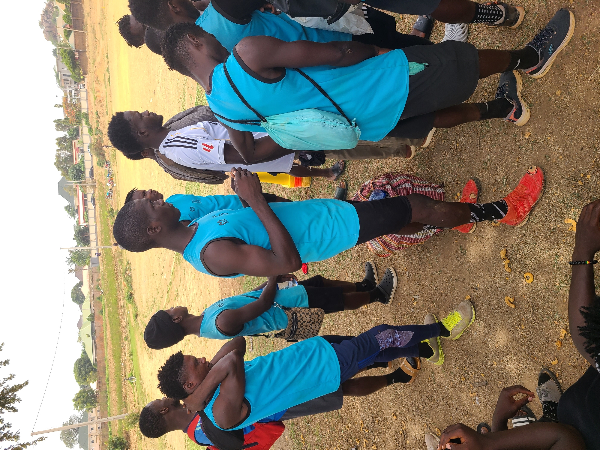 A group of young men is gathered in an open, grassy field in Barikin Jirga Summa, Nigeria. They are mostly wearing light blue athletic tank tops with black trim. Some are wearing shorts, while others have on athletic pants. Several of the individuals have water bottles and small bags or backpacks. They are standing and talking to each other. The field is a brownish color with sparse green grass. Some buildings and trees are visible in the background under a bright, overcast sky. The ground is littered with small, round objects.