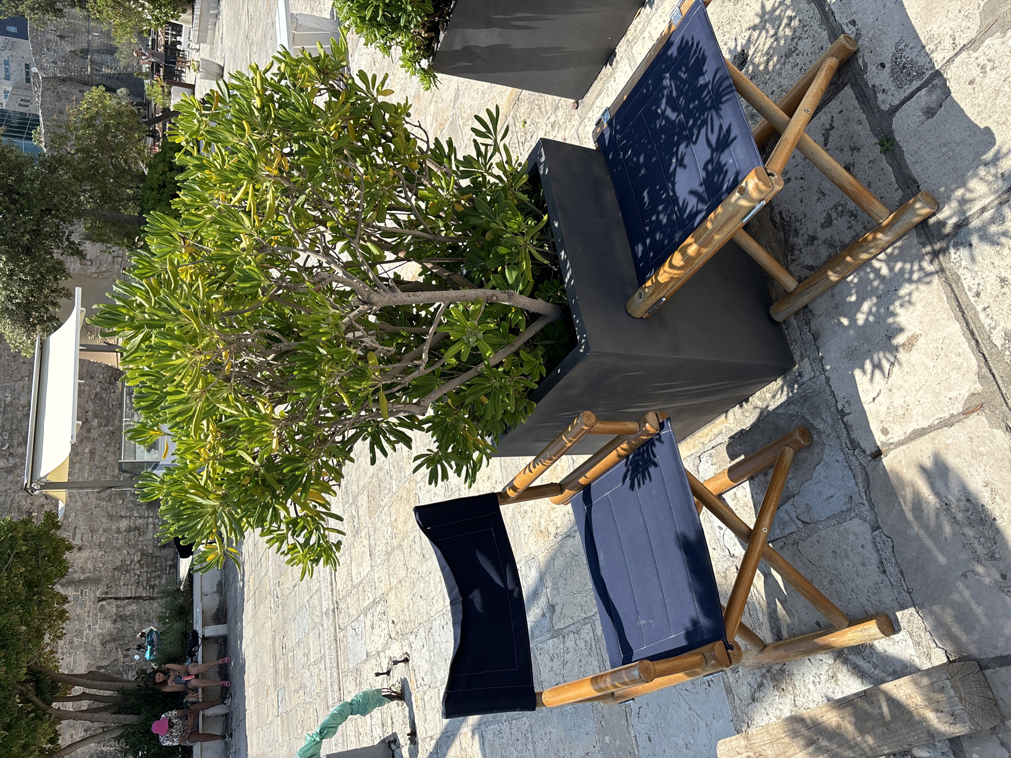 The image depicts an outdoor seating area in Budva, Montenegro. In the foreground, a dark blue director's chair with a wooden frame is positioned on a stone-paved ground. Beside it, a smaller, matching wooden stool with a dark blue seat is placed. Behind the chair and stool is a large, dark planter containing a lush green plant. In the background, several people are visible sitting on a stone bench under a tree. A motorcycle is parked behind them, and a stone wall forms the backdrop. Further in the distance, outdoor seating with tables and umbrellas suggests a cafe or restaurant setting. The overall environment is sunny, with dappled shadows from trees falling on the ground and furniture.