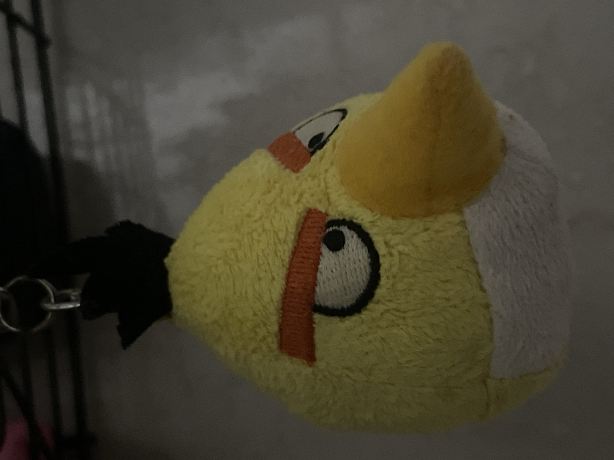 A close-up, side profile shot shows a yellow plush toy of the character Chuck from Angry Birds. The toy has a black tuft of hair on its head and a silver keychain attached to it. Its features, including its large eyes with black pupils and orange eyebrows, are embroidered onto the yellow fabric. A white patch is visible on its chin. The toy is hanging against a mottled grey background, possibly a wall. The lighting is dim, creating shadows and highlighting the texture of the plush material. There are no people or other objects clearly visible in the frame. The image does not contain any information that would allow for its location to be identified as Aktau, Kazakhstan.
