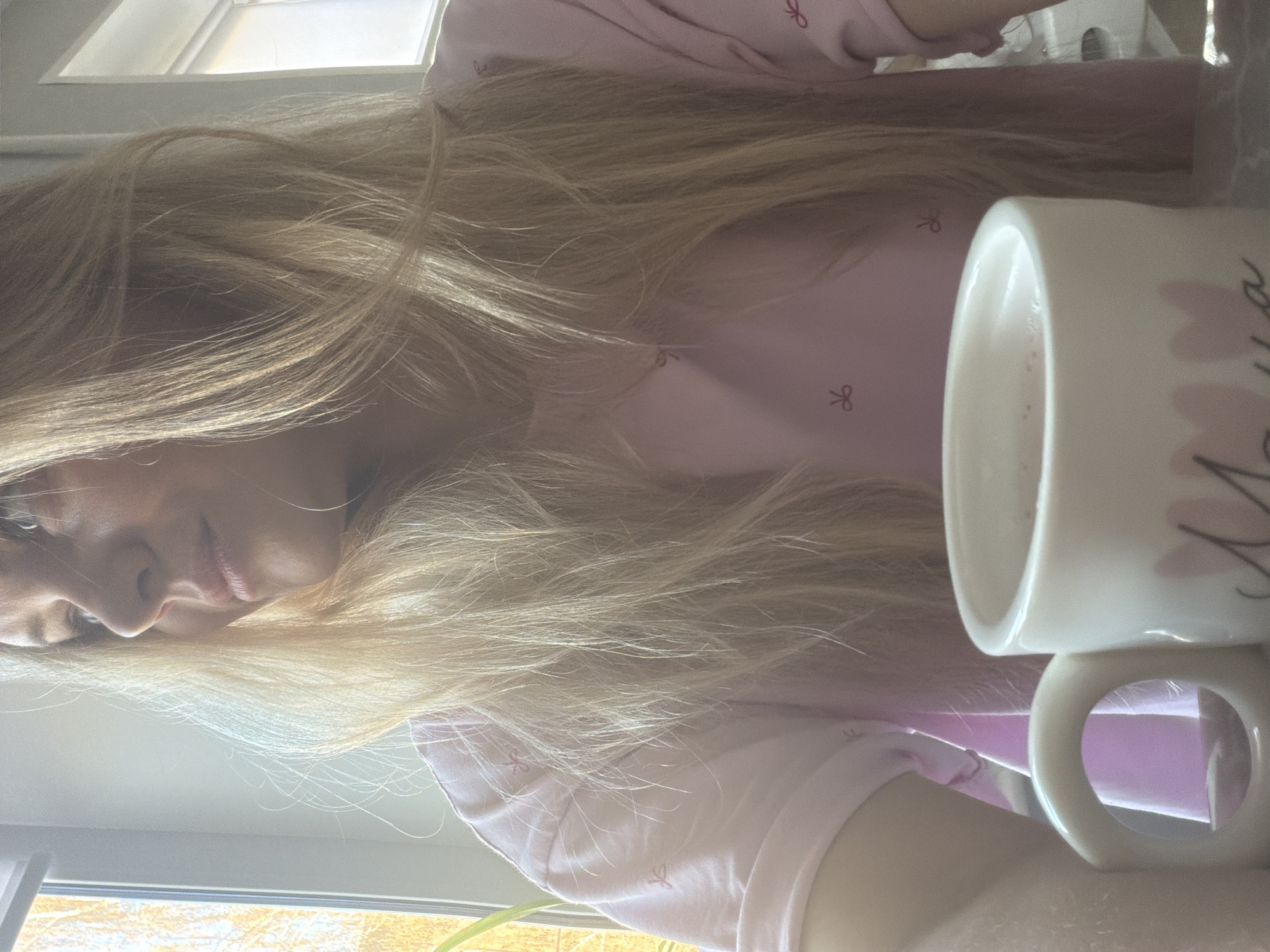 A fair-skinned person with long, blonde hair is positioned next to a window. The individual is wearing a light pink top with a bow pattern. A white mug, partly filled with a light-colored liquid, is visible near the person. The location is not discernible as Votkinsk, Russia. The setting appears to be indoors, illuminated by natural light filtering through the window.