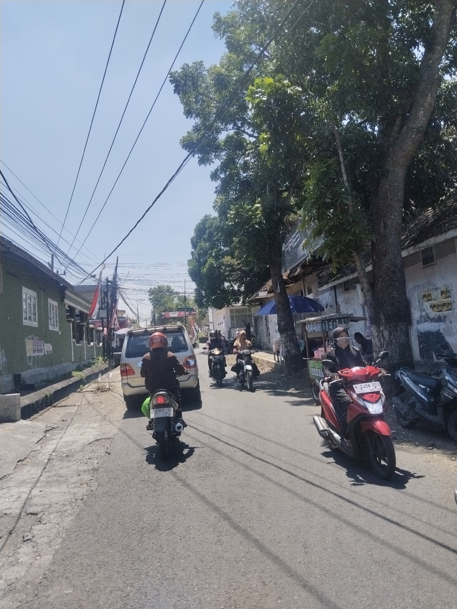 The image shows a street scene in Candirenggo, Indonesia. It is a sunny day with a clear sky. The street is paved and appears to be a road used by both cars and motorcycles. There are several trees lining the street, providing shade. On the left side of the street, there are buildings with green walls and white-framed windows. Some of these buildings have posters or signs on them.
In the foreground, a person on a motorcycle is riding away from the camera. The motorcycle is black with a green object attached to the back. The rider is wearing a dark jacket and a helmet. Another motorcycle is visible to the right, also being ridden by a person wearing dark clothing and a helmet. This motorcycle is red and has a license plate that reads "N 2458 DDO". There is a parked motorcycle to the far right of the frame.
Further down the street, there is a silver car with its rear facing the camera, and a person on a motorcycle with a red helmet is visible in front of it. More motorcycles are seen in the distance, as well as what appears to be a food cart. Power lines crisscross above the street and trees. The overall impression is of a typical urban street in Indonesia, with moderate traffic and local businesses.