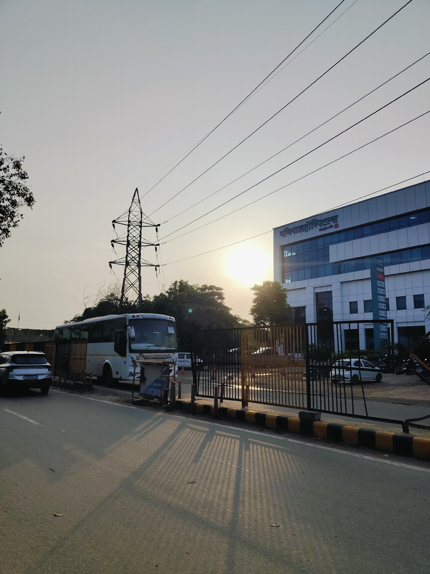 The image captures a view of Manipal Hospitals in Gurgaon, India, bathed in the warm glow of the setting sun. A white bus is parked on the side of the road, partially obscured by a temporary yellow barrier. To the left, a silver car is visible, its license plate reading "HR26CP0708". The hospital building, a modern structure with glass windows, stands prominently in the background. Overhead, power lines crisscross the sky, supported by a large electrical tower. A black metal gate separates the road from the hospital premises, and several vehicles, including a white car and motorcycles, are parked inside. The road in the foreground is asphalt, with yellow and black striped dividers along the edge. Shadows of the gate and power lines are cast across the road, indicating the time of day.