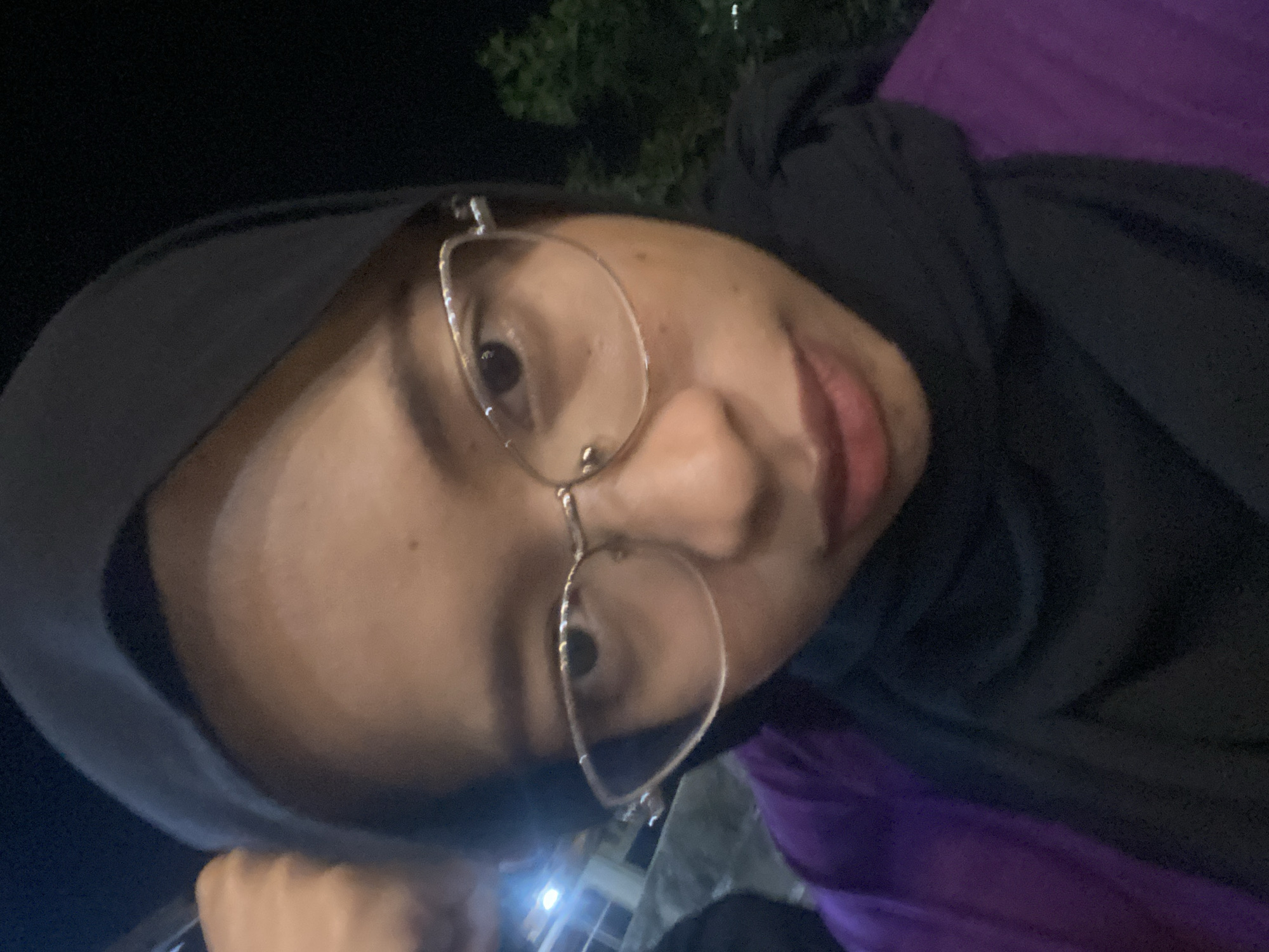 This is a close-up, head-and-shoulders selfie of a young woman wearing a black hijab and glasses. She is also wearing a purple garment over a black shirt. The image appears to have been taken at night, with some artificial lights visible in the background to the left, suggesting an outdoor urban or semi-urban setting. The woman's left arm is raised and her hand is resting on her head. Her expression is neutral, and she is looking directly at the camera. The background is dark, with some blurred greenery visible on the right side. The scene is likely in the city of Candirejo, Indonesia, given the provided context.