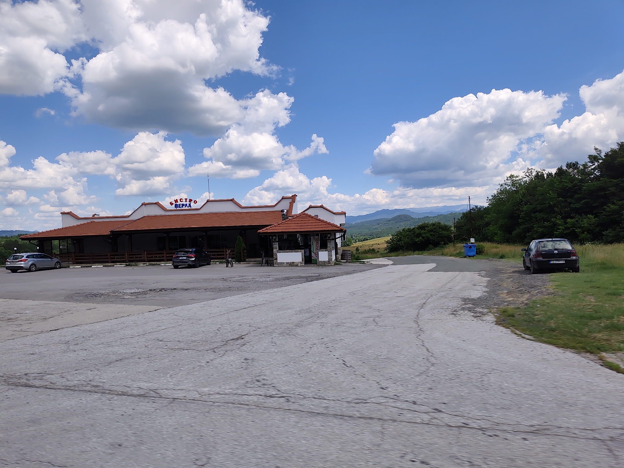This is a wide-angle, eye-level shot of a roadside establishment on a sunny day in Osikovitsa, Bulgaria. The building, with a red-tiled roof and white facade, appears to be a restaurant or cafe, with outdoor seating and a small kiosk. The name "БИСТРО ПЕРЛА" (BISTRO PERLA) is visible in blue lettering on the building's facade.
The scene is set against a backdrop of rolling green hills and mountains under a bright blue sky dotted with fluffy white clouds. The foreground consists of a cracked asphalt parking lot and road, suggesting a well-trafficked area. Several cars are parked in front of the establishment, including a silver SUV and a dark-colored sedan. A blue trash bin is also visible.
The lighting suggests it is daytime, likely mid-morning or afternoon, with ample sunlight casting shadows. The overall atmosphere is one of a typical rural stopover, offering a place to rest and refuel amidst scenic Bulgarian landscape. There are no people prominently featured, though a few figures are vaguely discernible near the building entrance, their activities unclear. The weather appears pleasant, suitable for outdoor activities.