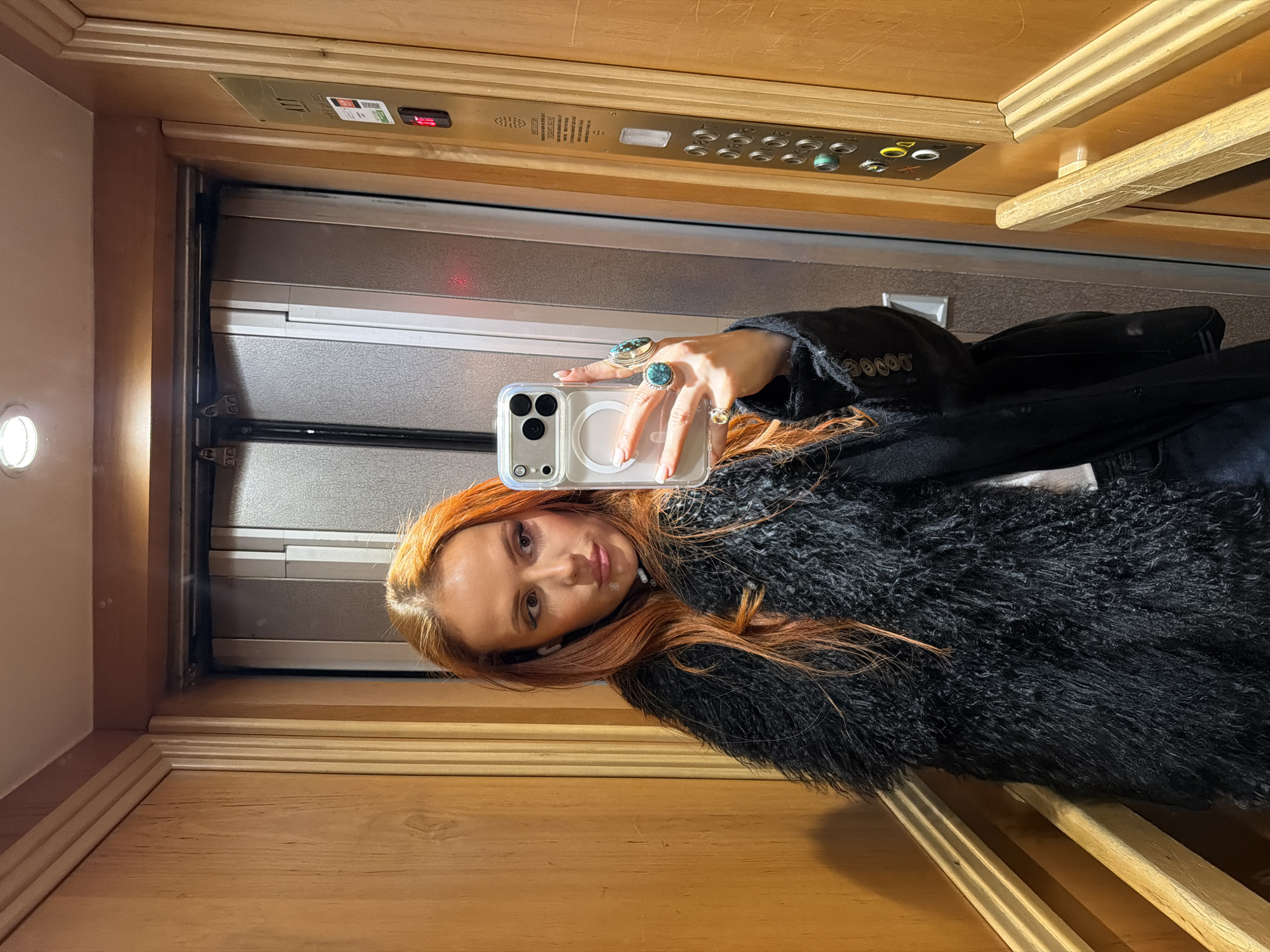 A female subject with long, reddish-orange hair is taking a mirror selfie inside an elevator within the city of Paris, France. She is wearing a black, shaggy faux-fur coat over a dark garment. Her right hand, adorned with multiple large turquoise rings and a smaller gold-toned ring, holds a silver-colored smartphone in a clear MagSafe-compatible case, positioned to capture her reflection. White nail polish is visible, and white earbuds are present in both ears. The subject looks directly into the phone's lens with a slight pout.
The elevator interior features light-toned wooden paneling on the walls, characterized by vertical fluting and a horizontal wooden handrail on the right. The elevator doors, visible behind the phone, are constructed from textured, light-grey metallic panels. On the right wall, a metallic control panel displays a red digital "1," indicating the current floor. Below this, unreadable French text is present, followed by a vertical array of circular buttons, including illuminated green, yellow, and red indicators, along with door control symbols. A single circular recessed light illuminates the ceiling.
