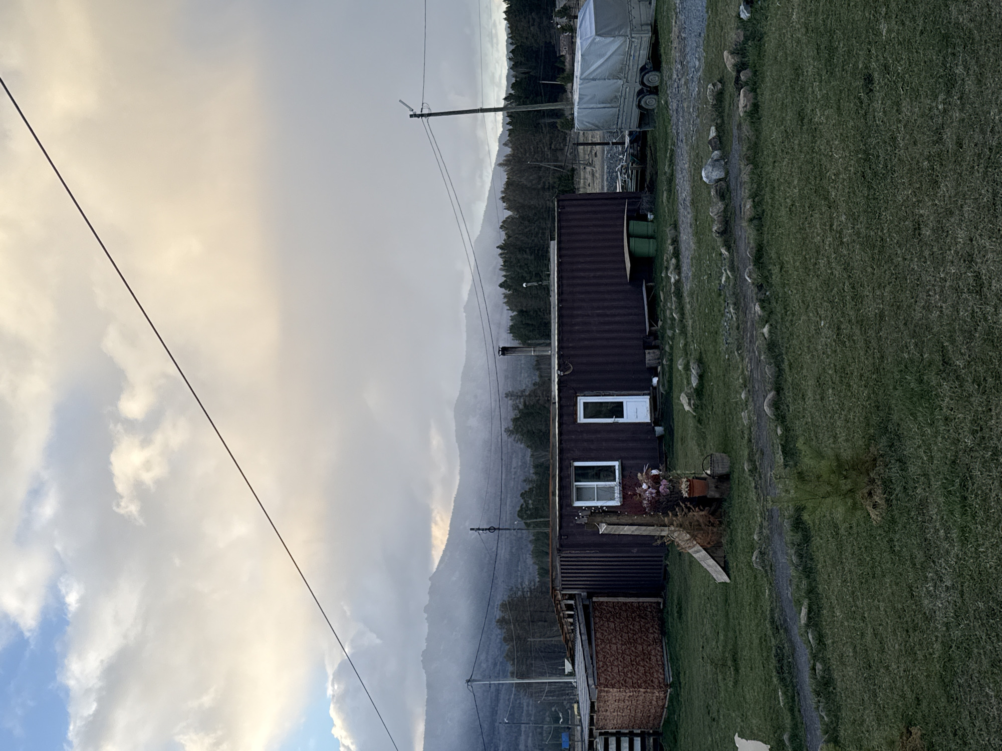 The image depicts a rural scene near Tyulyuk, Russia. A dark, two-story container home sits adjacent to a large grassy area, bordered by a rocky path. A trailer is parked further up the path. Mountains loom in the background, partially obscured by clouds. Utility lines are visible in the sky.