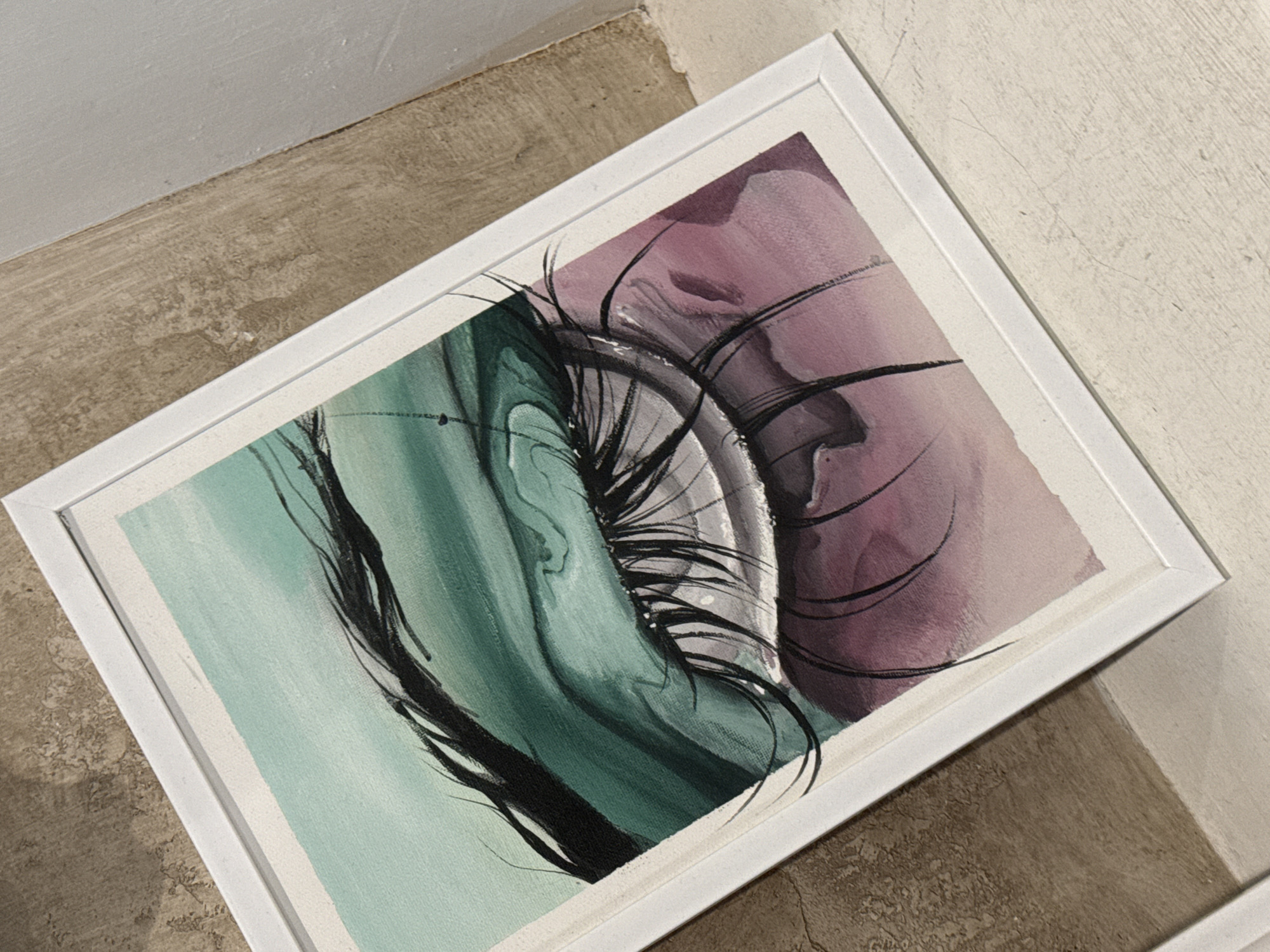 The image displays a framed abstract painting tilted on a textured concrete floor, possibly indoors. The painting features vibrant streaks of teal and a soft purple, blended with dark, gestural brushstrokes that suggest an eye with dramatic eyelashes. The framing is simple and white, and the canvas shows a textured surface. The lighting suggests it is daytime, casting soft shadows. The setting appears to be an unfinished or industrial-like space, given the rough concrete floor and the unpainted wall. No text is visible in the image. The artwork itself evokes a sense of artistic expression and perhaps a touch of mystery or intensity through its abstract representation.