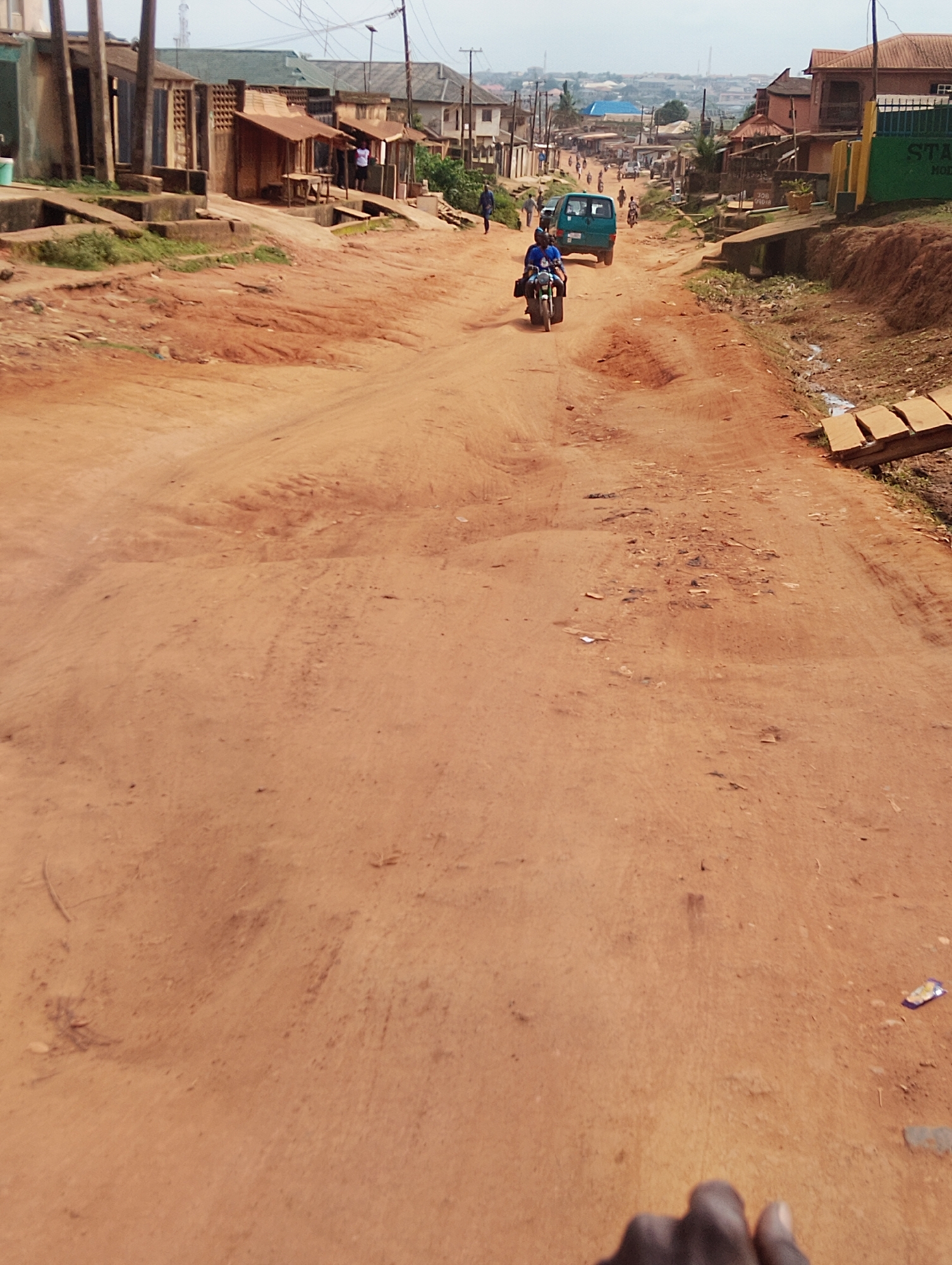 The image depicts a wide, unpaved dirt road in a developing urban area, likely Agege, Nigeria. The road is a reddish-brown color, indicating it is likely composed of laterite soil, and shows signs of wear and tear with ruts and uneven surfaces. Buildings line both sides of the road, appearing to be a mix of residential and commercial structures, some with corrugated iron roofs and others with more permanent construction. These buildings are modest and appear to be close to the roadside.
In the middle of the road, a turquoise blue van is traveling away from the viewer. Ahead of the van, a person is riding a motorcycle, also moving away from the viewer. The motorcycle rider appears to be wearing a blue shirt and possibly carrying a bag. Other individuals are visible in the background, walking along the sides of the road and near the buildings, suggesting a normal day of activity. The sky is overcast, contributing to the muted lighting of the scene. On the right side of the road, there is a distinct drainage channel, and a few wooden planks are placed across it, serving as a makeshift bridge or walkway. The overall impression is one of a bustling, but informal, urban environment.