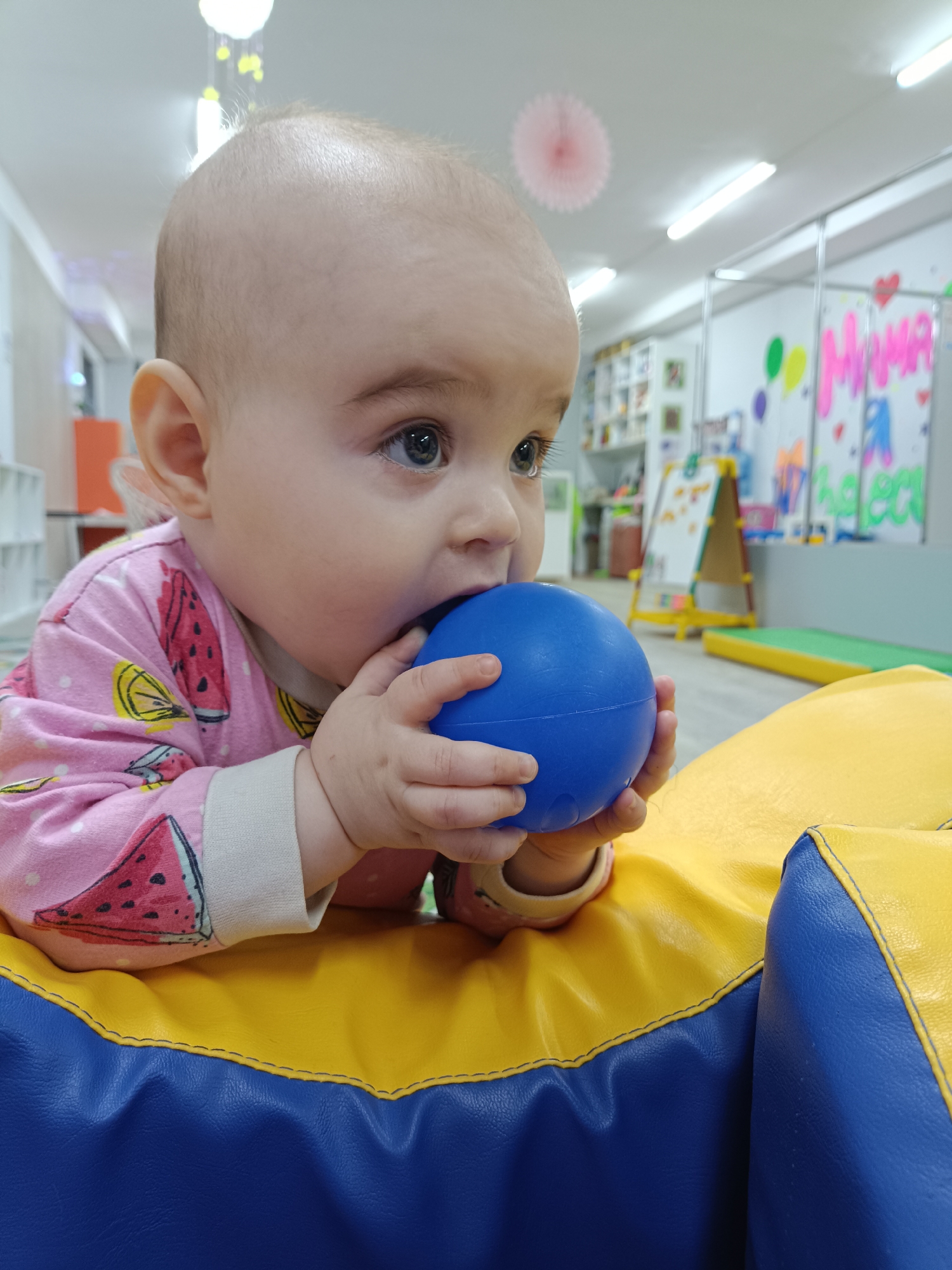 A baby is lying on a yellow and blue cushion, with a blue ball in their mouth and held by both hands. The baby is wearing a pink pajama top with watermelon and lemon designs. The location appears to be an indoor play area, with a whiteboard, toys, and various colorful decorations in the background. The city is unknown.