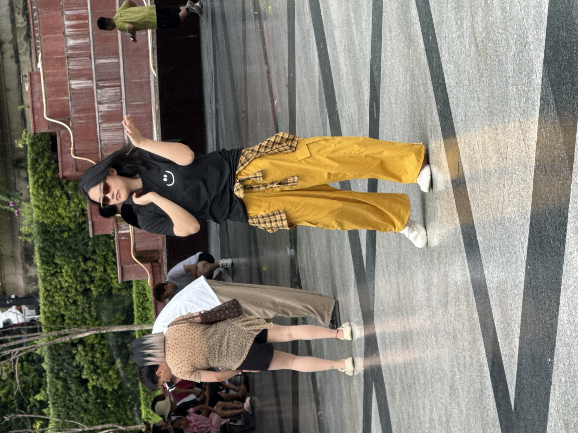 In Yuzhong District, China, a young woman stands on a patterned gray tiled plaza. She is wearing a black t-shirt with a white smiley face logo, loose mustard-yellow cargo pants, and white sneakers. A plaid shirt, yellow, brown, and black, is tied around her waist. She has sunglasses on and her left hand is raised, palm open, as if gesturing or posing. Behind her, other people are visible, including a person in a light-colored shirt and brown pants, and further back, a child in a green shirt and black shorts. The background features tiered red structures and lush green bushes. The ground has a reflective quality, showing distorted light patterns.