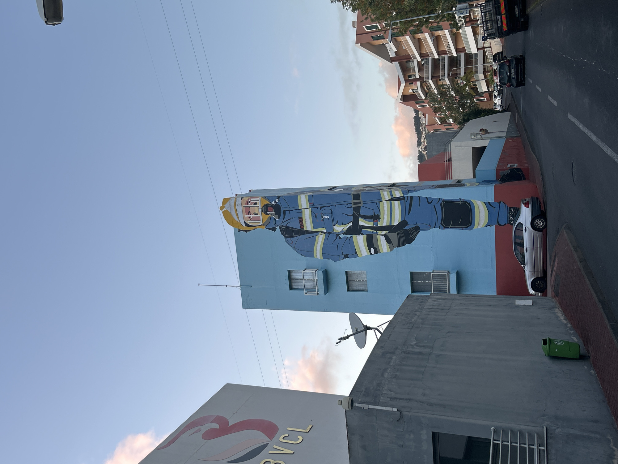 The image shows a street in Câmara de Lobos, Portugal, featuring a tall building with a mural of a firefighter on its side. The mural depicts a figure in full firefighter gear, rendered in a cartoonish style, against a blue background. The building itself is partially blue and red. The street is lined with other buildings and parked cars. A green trash bin sits against a gray wall in the foreground. Overhead, power lines crisscross the sky. A company sign marked BVCL is also visible.