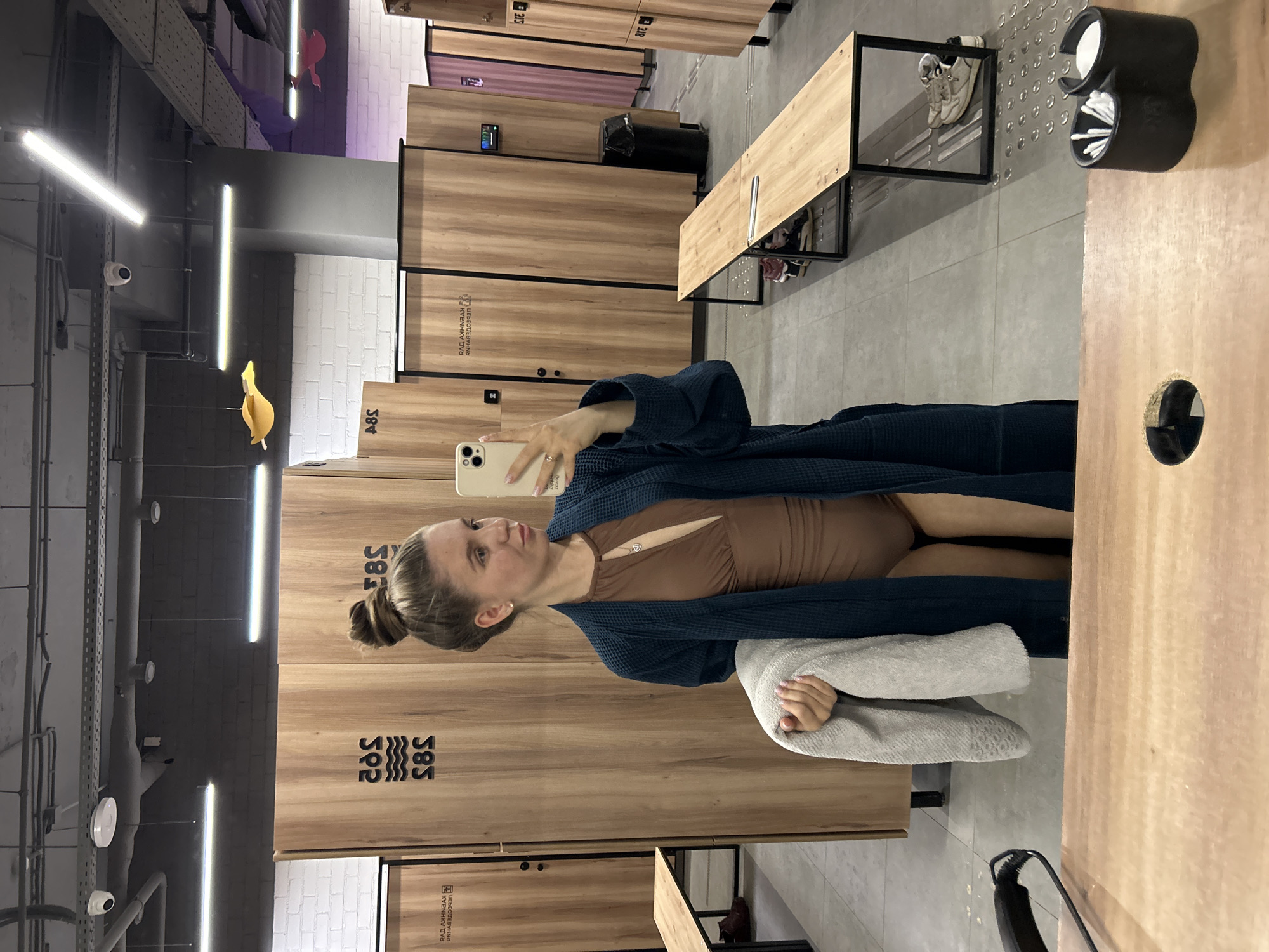 A young woman is captured in a mirror selfie within what appears to be a modern changing room or locker facility. The image is oriented sideways, with the woman standing upright, holding a light-colored smartphone to take the photo. Her light brown hair is pulled back in a bun. She wears a dark blue, waffle-knit robe over a light brown one-piece garment, possibly a swimsuit or bodysuit, and holds a light gray towel in her left arm.
The setting features rows of light wood-grained lockers, some marked with black numbers like "585" and "582," and a stylized wave logo. Upside-down text on a locker door reads "РАДИ ДВИЖЕНИЯ К УСПЕХУ" (FOR MOVEMENT TOWARDS SUCCESS). The floor is covered with gray square tiles, and the walls are a mix of white brick and dark gray. Overhead, exposed industrial-style piping and bright, elongated LED strip lights illuminate the space. A wooden bench with a black metal frame stands to the right, with a pair of sneakers visible beneath it. A small black security camera is mounted on the left wall/ceiling. Also visible is a yellow, duck-shaped decorative item hanging from the ceiling and a black wall-mounted dispenser containing cotton pads and swabs. The overall ambiance is clean and functional, suggesting a gym, spa, or aquatic center changing area.