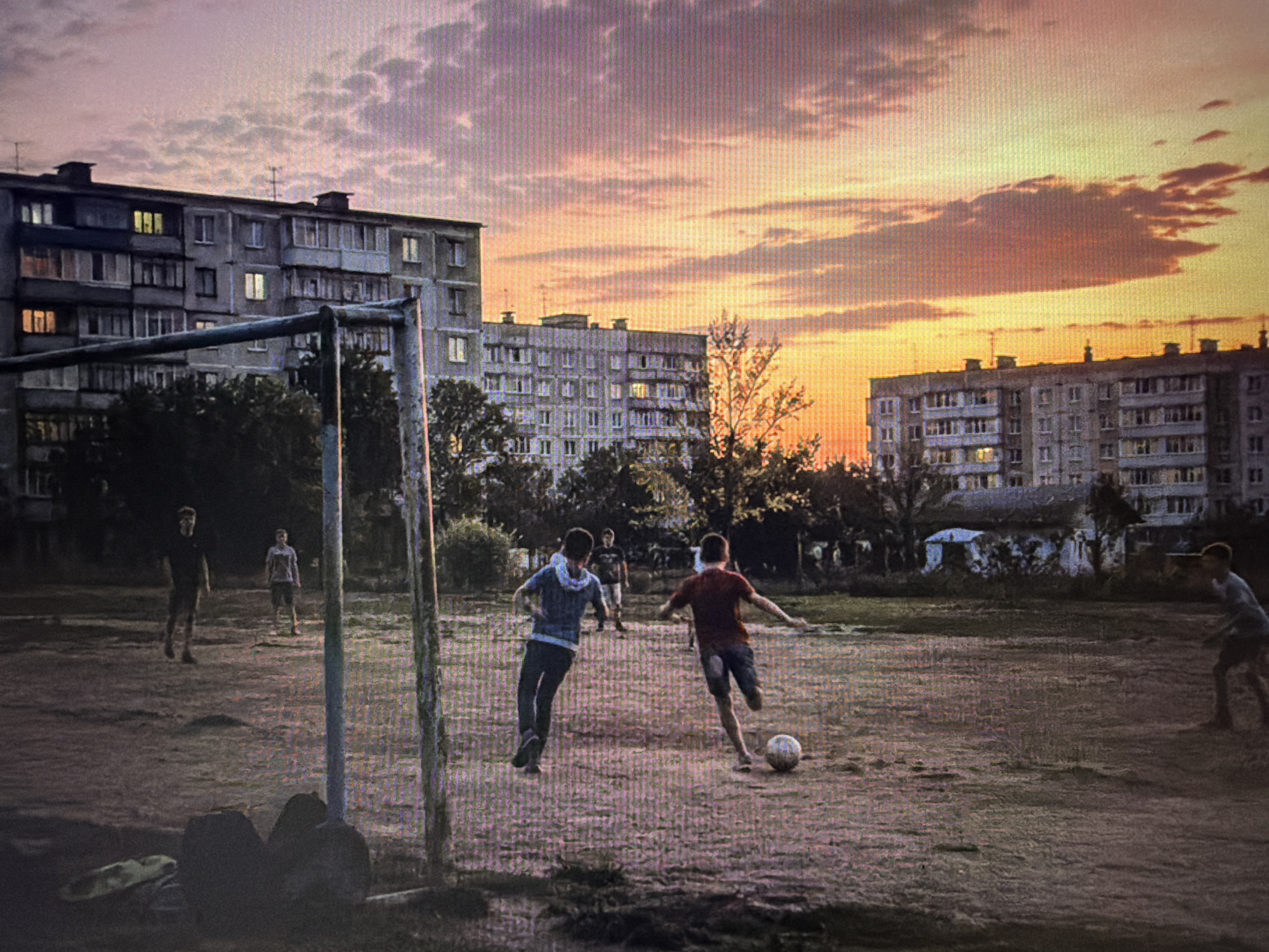 This image captures a group of young people playing soccer on a dusty field at dusk. The sky is a dramatic mix of pink, orange, and purple clouds, illuminated by the setting sun. In the background, several multi-story apartment buildings, characteristic of Eastern European urban landscapes, are visible. Some windows in the buildings are lit, suggesting that people are home.
The focus of the image is the soccer game. Two figures are prominently in the foreground, actively engaged with the ball. One player, wearing a red shirt and dark shorts, is mid-kick, while another, in a blue hoodie and dark pants, is positioned defensively. Two other players are visible further back in the field, seemingly observing or waiting for their turn. A soccer goal, weathered and slightly dilapidated, stands to the left of the frame, further emphasizing the casual, local nature of the game.
The overall mood is one of youthful energy and simple enjoyment, set against a backdrop of urban infrastructure and a beautiful, fading sky. The time of day is clearly evening, with the fading light casting long shadows and lending a somewhat muted, atmospheric quality to the scene. The ground is dry and uneven, indicating a well-used, possibly neglected, playing area. There is no visible text in the image.