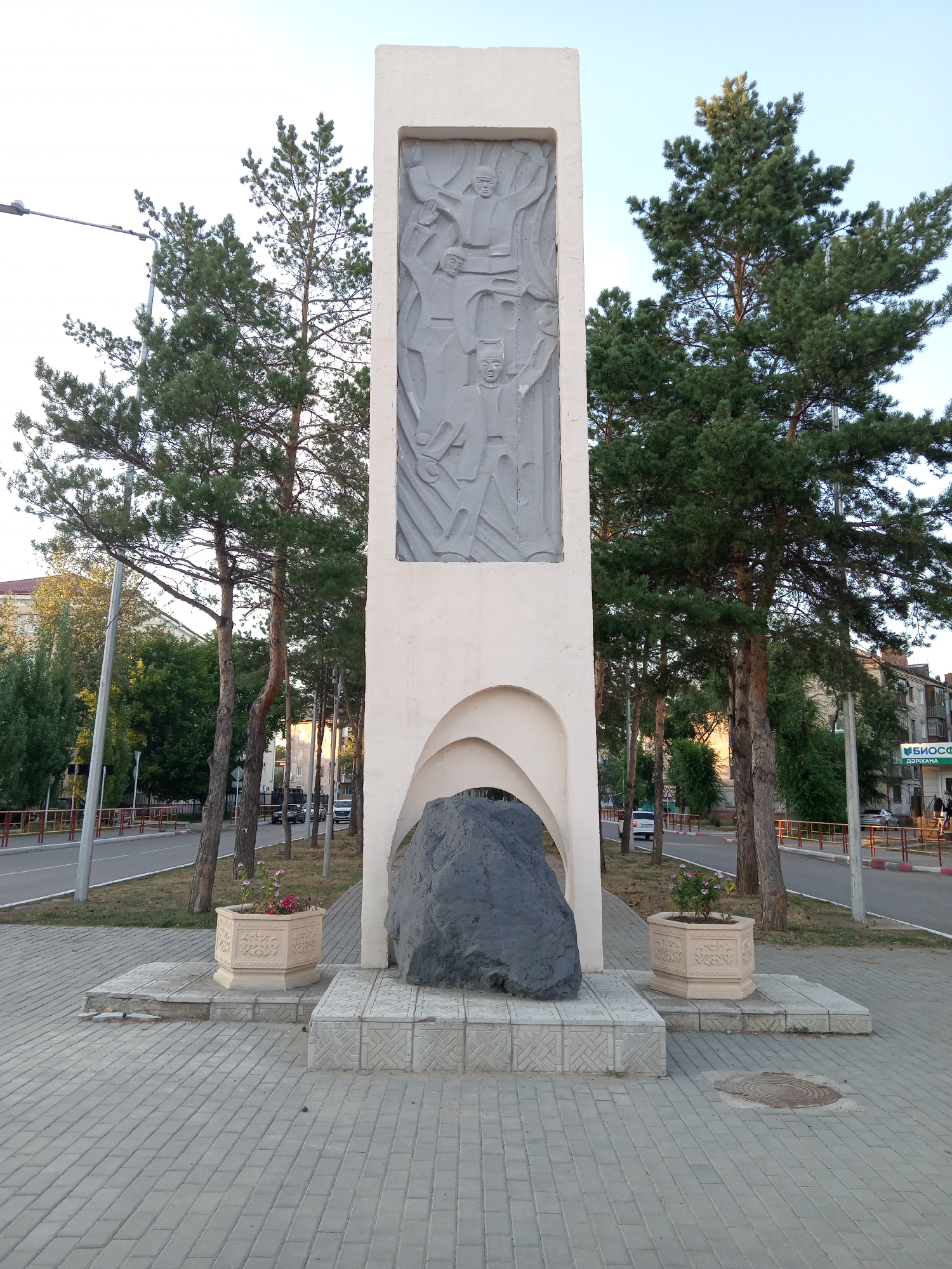 The image shows a monument in the city of Rudny, Kazakhstan. The monument is a tall, rectangular structure with a bas-relief on its upper half depicting several figures. At the base of the monument is a large, dark gray boulder. Flanking the boulder are two hexagonal planters containing small pink flowers. The monument stands on a paved area made of gray interlocking bricks. Behind the monument, tall pine trees are visible, and beyond them, buildings and a road. The sky is clear and blue, suggesting daytime. The overall scene is a public space, possibly a park or a memorial area, in a city environment.