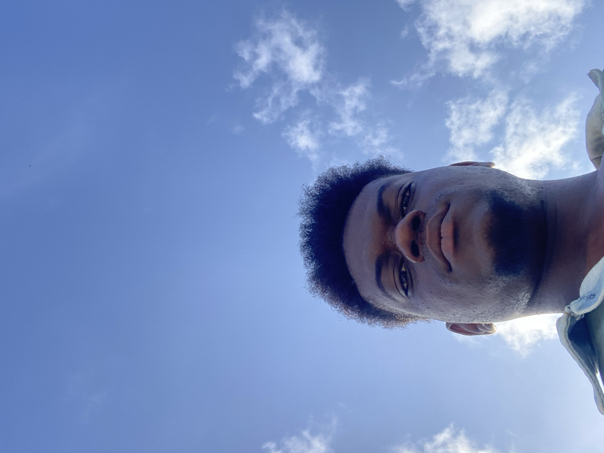 A low-angle shot captures a man against a backdrop of a blue sky dotted with clouds. The man's face is the primary subject, positioned at the top right of the frame. His skin tone is dark, and he has short, curly hair. A beard partially covers his jawline. His expression is neutral. The sky is a gradient of blue, with patches of white clouds scattered throughout. The location is undetermined.