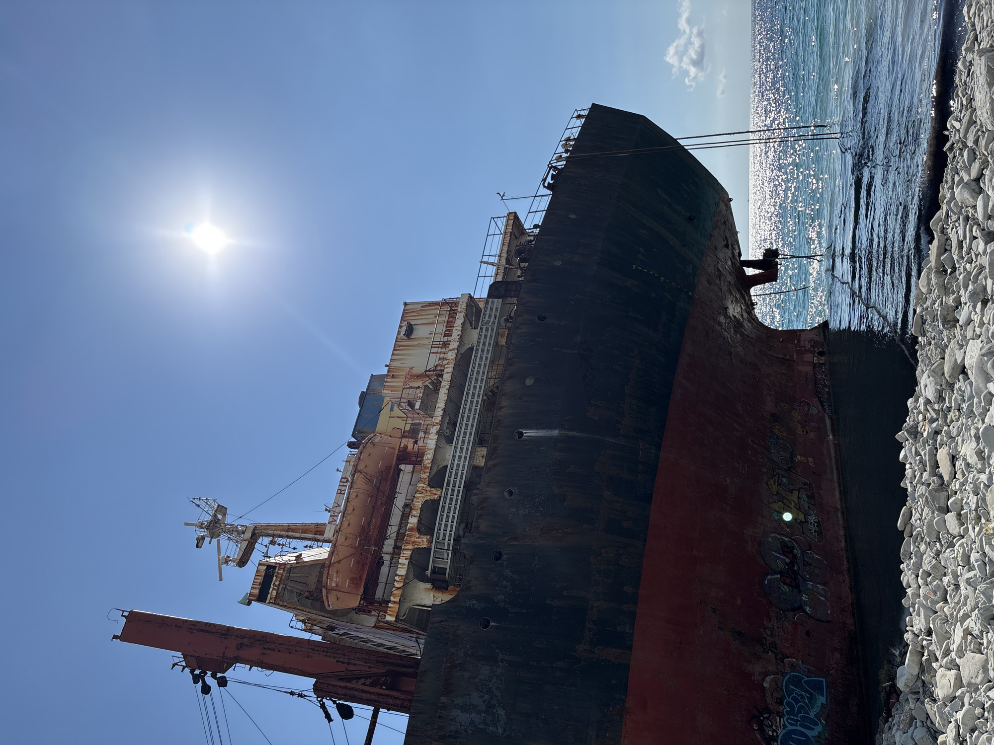 The image depicts the abandoned ship MV Rio, beached on the shore in Kabardinka, Russia. The ship is heavily rusted and appears to be in a state of disrepair. The hull is partially submerged in the clear blue water, and the deck is littered with debris. The sun is shining brightly in a clear blue sky, casting a glare on the water. The shore is covered with white pebbles. The overall scene suggests a sense of decay and neglect, with the ship serving as a stark reminder of its former glory.