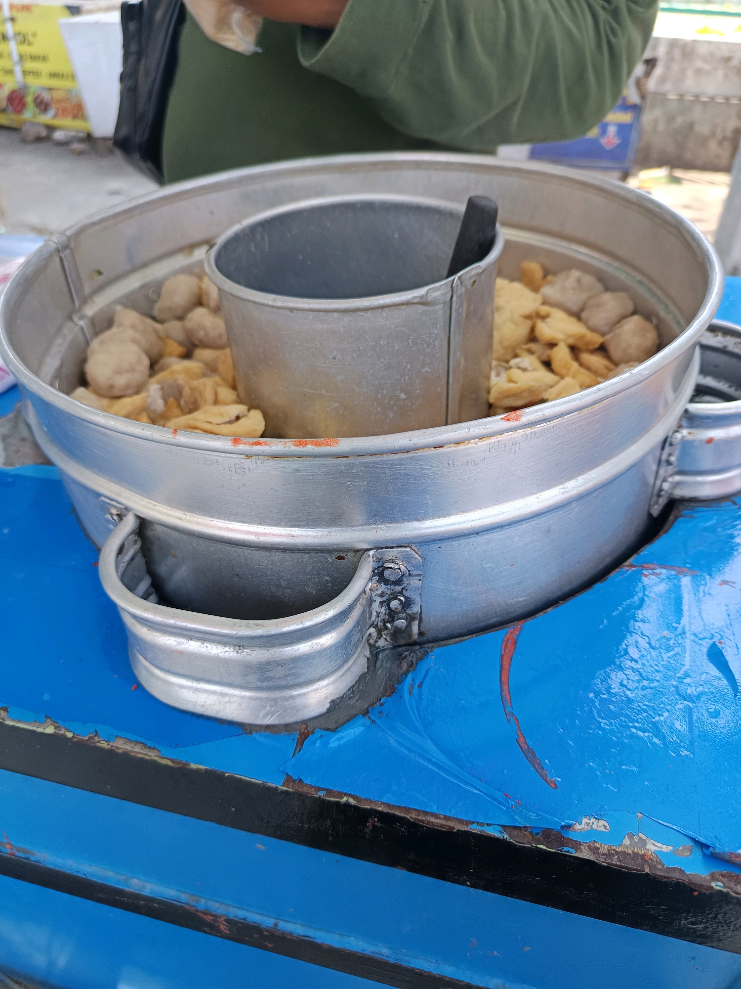 This is a close-up, eye-level, outdoor shot of a food stall in Sampang, Indonesia. The primary subject is a large, aluminum food steamer containing what appears to be meatballs and fried tofu. The steamer has two visible compartments, with the inner compartment holding the food and a smaller, removable metal cup with a black utensil. The steamer is placed on a blue surface, likely a table or cart, which shows signs of wear and peeling paint. In the background, a person wearing a green garment and a black bag is partially visible, along with a yellow sign that is out of focus. The overall lighting suggests it is daytime.