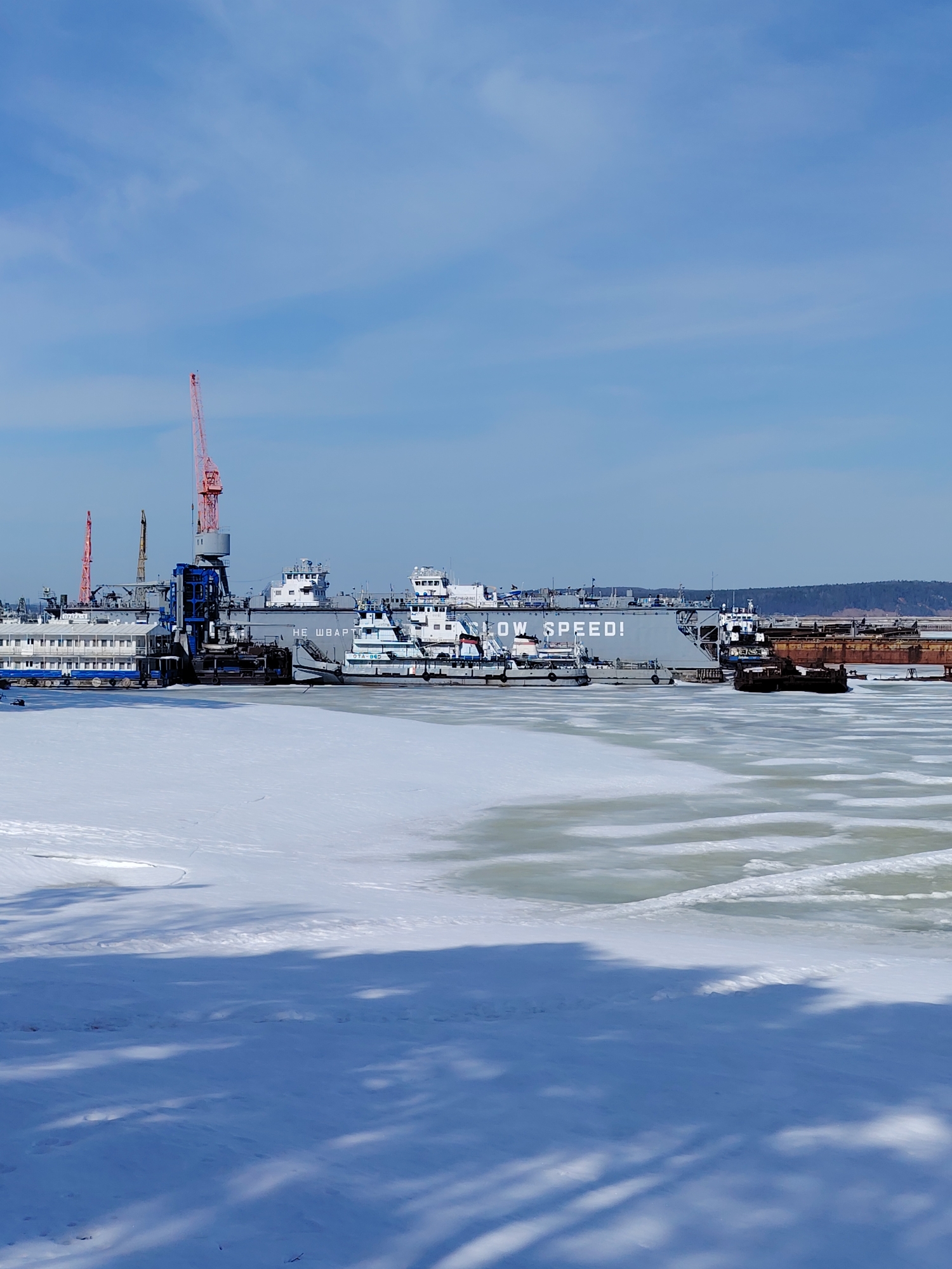 The image depicts a winter scene in Chaikovsky, Russia, showing a partly frozen body of water in the foreground and various ships and cranes docked along the shore in the background. A large gray barge is visible with the phrase "SLOW SPEED!" printed on its side, alongside smaller tugboats and a floating dock structure. Three red cranes stand amongst the ships and docking structures. The foreground is covered in snow and ice, with visible shadows of trees or structures. The sky is light blue with some scattered clouds. The overall scene suggests an industrial harbor area during winter.