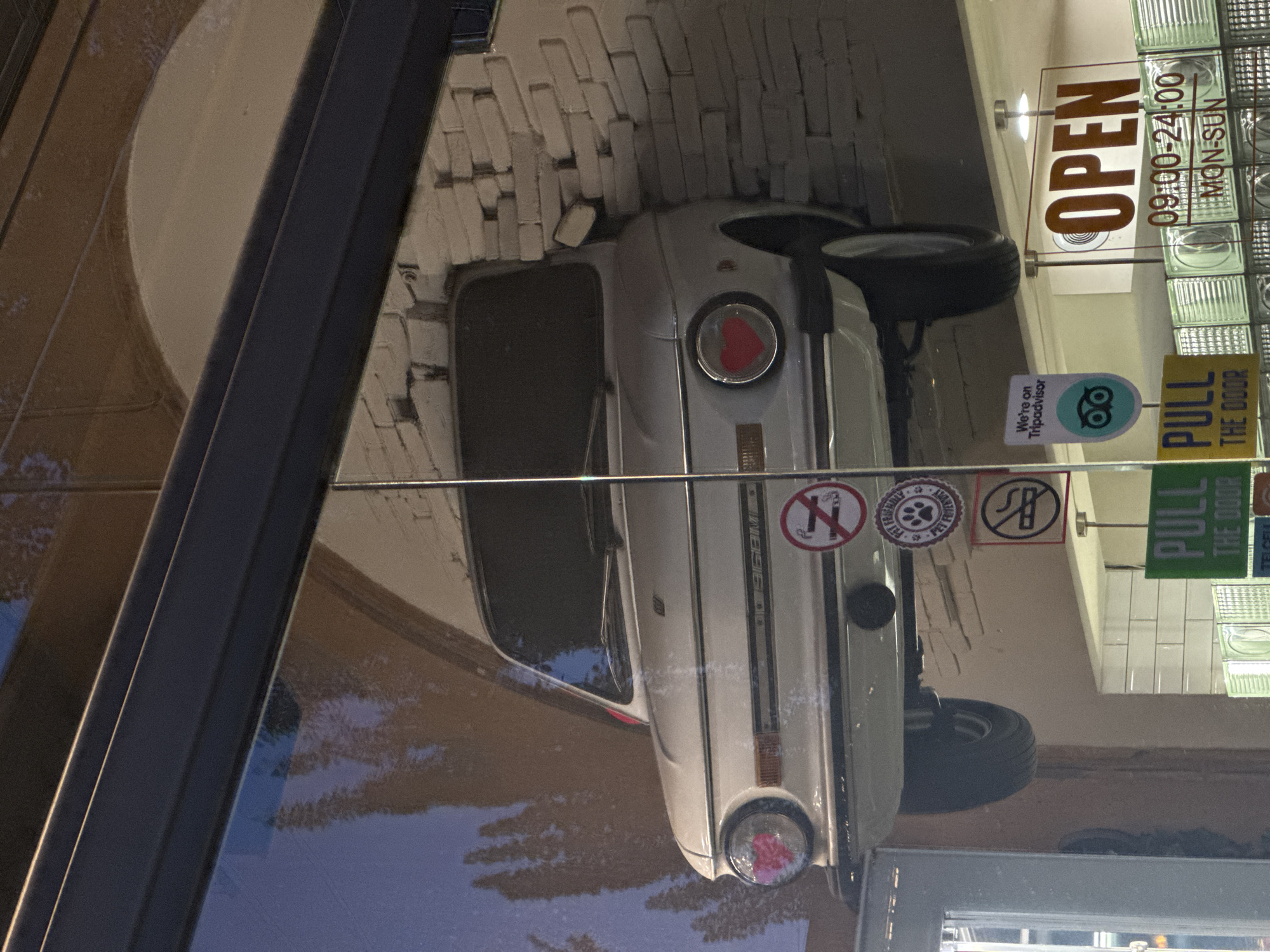 A still image shows a reflection of a store front in Yerevan, Armenia. Inside is a white vintage car mounted on the ceiling, with a red heart on one of the headlights. There are signs on the window, including one that says "OPEN 09:00-24:00, MON-SUN", and others displaying no smoking, pet friendly and no bikes allowed symbols. Another sign says "Pull the door". The bricks above the car are painted white, and the glass block wall is in the upper right corner of the image.