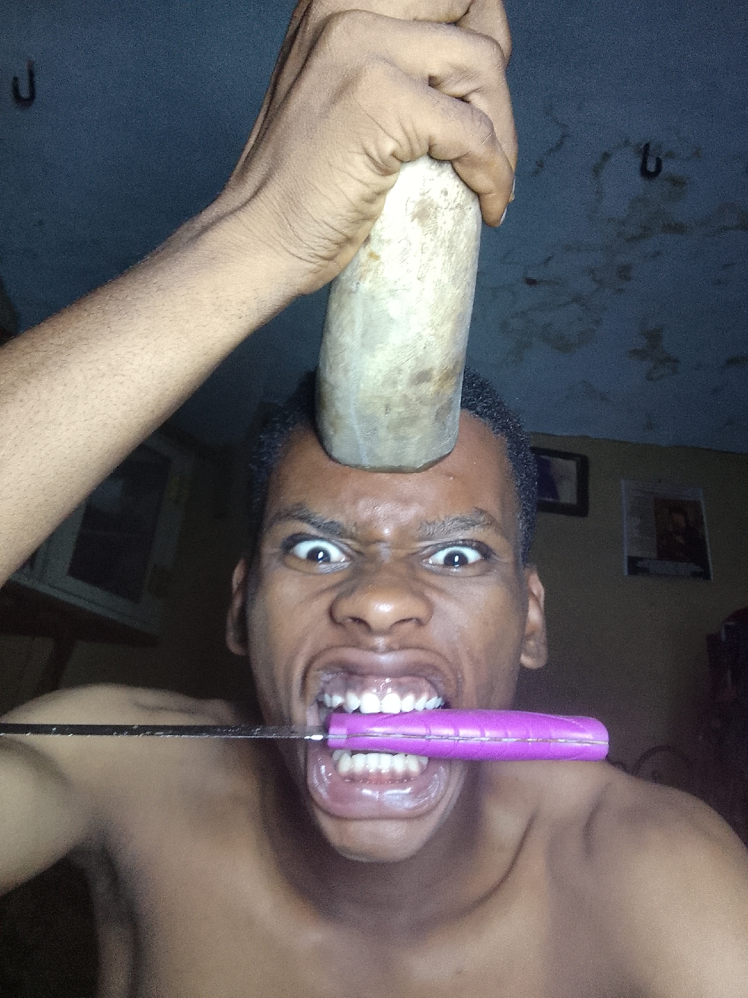 A close-up shot depicts a dark-skinned man with close-cropped hair, expressing a strained facial expression with widened eyes and bared teeth. A cylindrical object, likely made of wood or a similar material, is pressed against the center of his forehead, held in place by a hand. A flat object with a purple handle is held horizontally in his mouth. The setting appears to be indoors with hooks visible on the ceiling and two framed pictures in the background. The location is Unknown, Unknown.