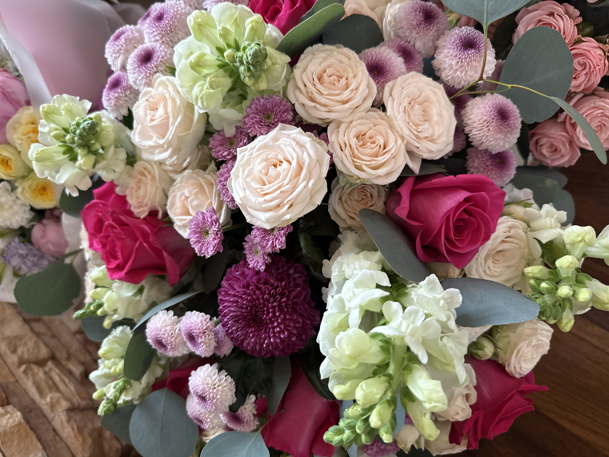 This media file is a close-up, top-down shot of a vibrant floral arrangement. The bouquet features a variety of flowers in shades of pink, cream, white, and deep fuchsia, interspersed with delicate green foliage. Prominent among the blooms are roses with tightly packed petals, resembling miniature peonies, in soft cream and blush tones. There are also clusters of small, spherical purple and pink chrysanthemums, some with lighter tips and darker centers, adding texture and contrast. White snapdragons or similar flowers with multiple buds are also visible, some in a pale green hue. A few dark pink or fuchsia roses are nestled within the arrangement, providing a bold splash of color. The overall impression is one of a lush and romantic bouquet. The background is divided, with the left side showing a textured surface that appears to be stacked stones or bricks, while the right side is a darker, smooth surface, likely a wooden table. There is no text visible in the image, nor are there any people. The lighting suggests it is daytime, likely indoors given the focus on the floral arrangement. The style of the bouquet hints at a special occasion, possibly a gift or decoration for an event. The location context provided is Votkinsk, Russia, but there are no specific visual cues within the image to confirm this.