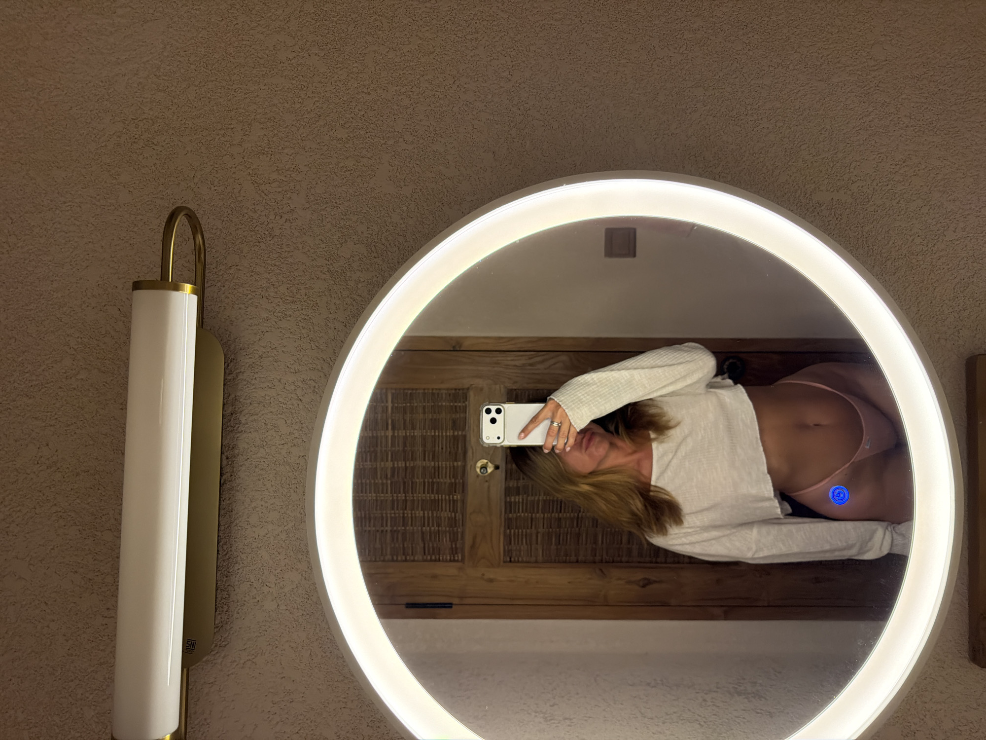 An eye-level mirror selfie depicts an individual in a bathroom setting in Mengwi, Indonesia. The person, with long brown hair, wears a white long-sleeved cropped top revealing their midriff and pink string-bikini style underwear. They hold a white iPhone in a clear case with their right hand, obscuring their face. A silver ring is visible on the ring finger of the right hand.
The reflection is contained within a circular mirror with an illuminated white LED rim, featuring a blue circular touch sensor on the lower left portion of the mirrored surface. Reflected behind the individual is a wooden door with a woven rattan panel in its upper section, featuring a black hinge and brass-colored door hardware including a keyhole plate and handle. A portion of a white terrycloth garment, possibly a bathrobe or towel, is also reflected on the left side of the mirror.
Above the mirror, a white cylindrical light fixture with brass accents is mounted horizontally on a textured, light brown stucco wall. The wall texture is consistent throughout the visible background outside the mirror's reflection.
