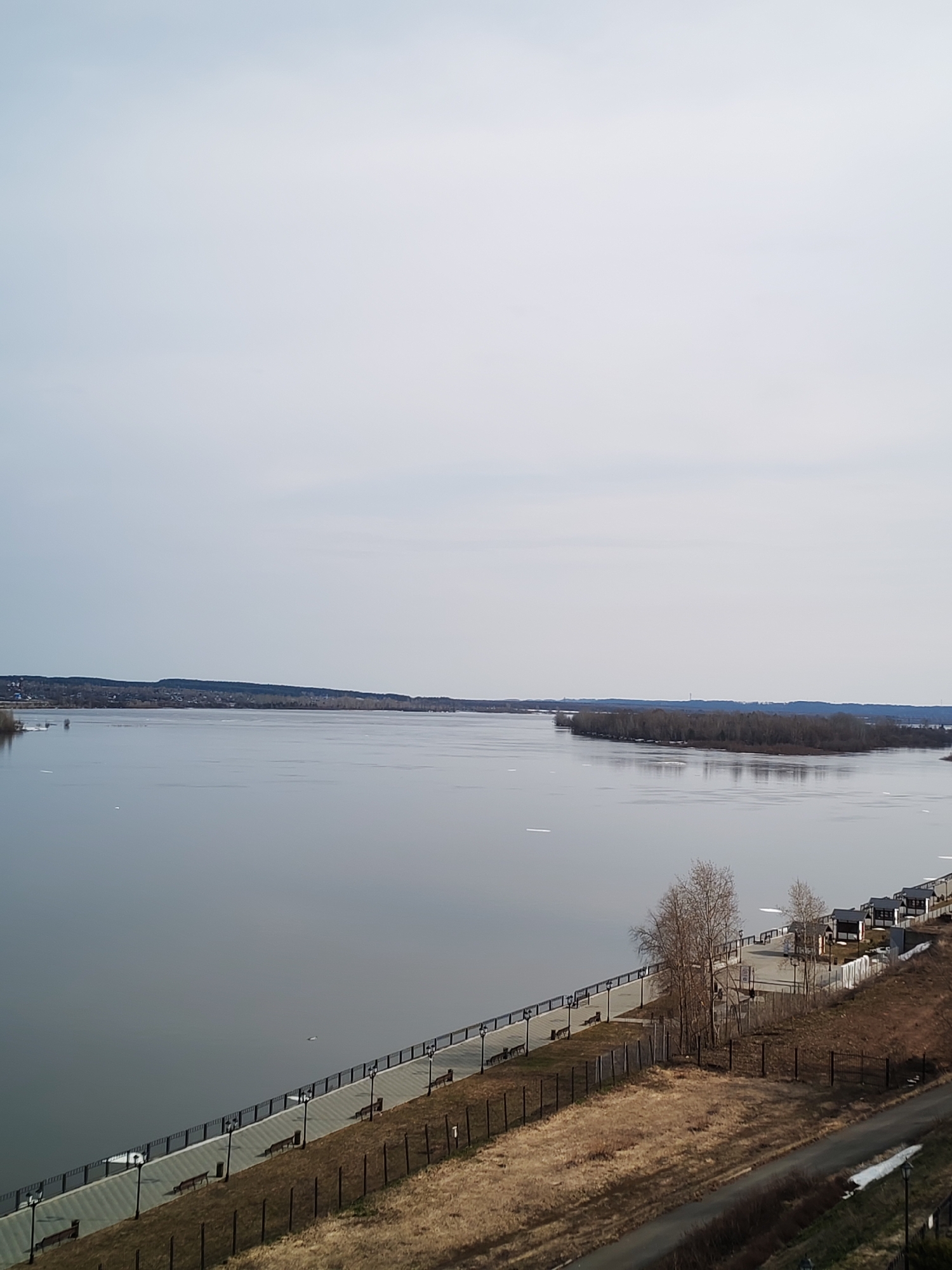 The image depicts a panoramic view of the Kama River in Sarapul, Russia. The river is wide and calm, reflecting the overcast sky. Small ice floes are scattered on the water's surface. An island covered in trees is visible in the middle of the river.
A paved promenade runs along the riverbank. Benches and lampposts are spaced along the promenade. A grassy slope descends from the promenade towards a lower path. A fence runs along the edge of the slope. Small buildings and structures are visible along the promenade.