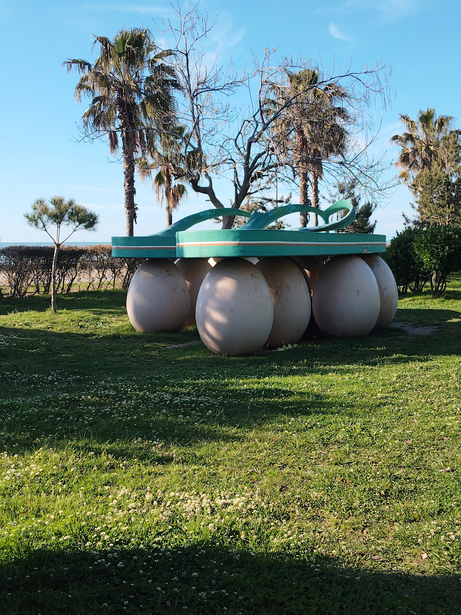 The image shows a sunny day in a park-like setting, likely in Batumi, Georgia. In the foreground, there is an art installation that consists of a large, green and white sculpture of a flip-flop sandal balanced atop a collection of six large, pale, spherical objects. The installation is positioned on a patch of green grass scattered with small white flowers. Behind the sculpture, there are several palm trees and other trees with some bare branches, under a clear blue sky. In the distance, there is a hint of a beach. The shadows cast on the grass indicate the direction of the sun and the time of day.