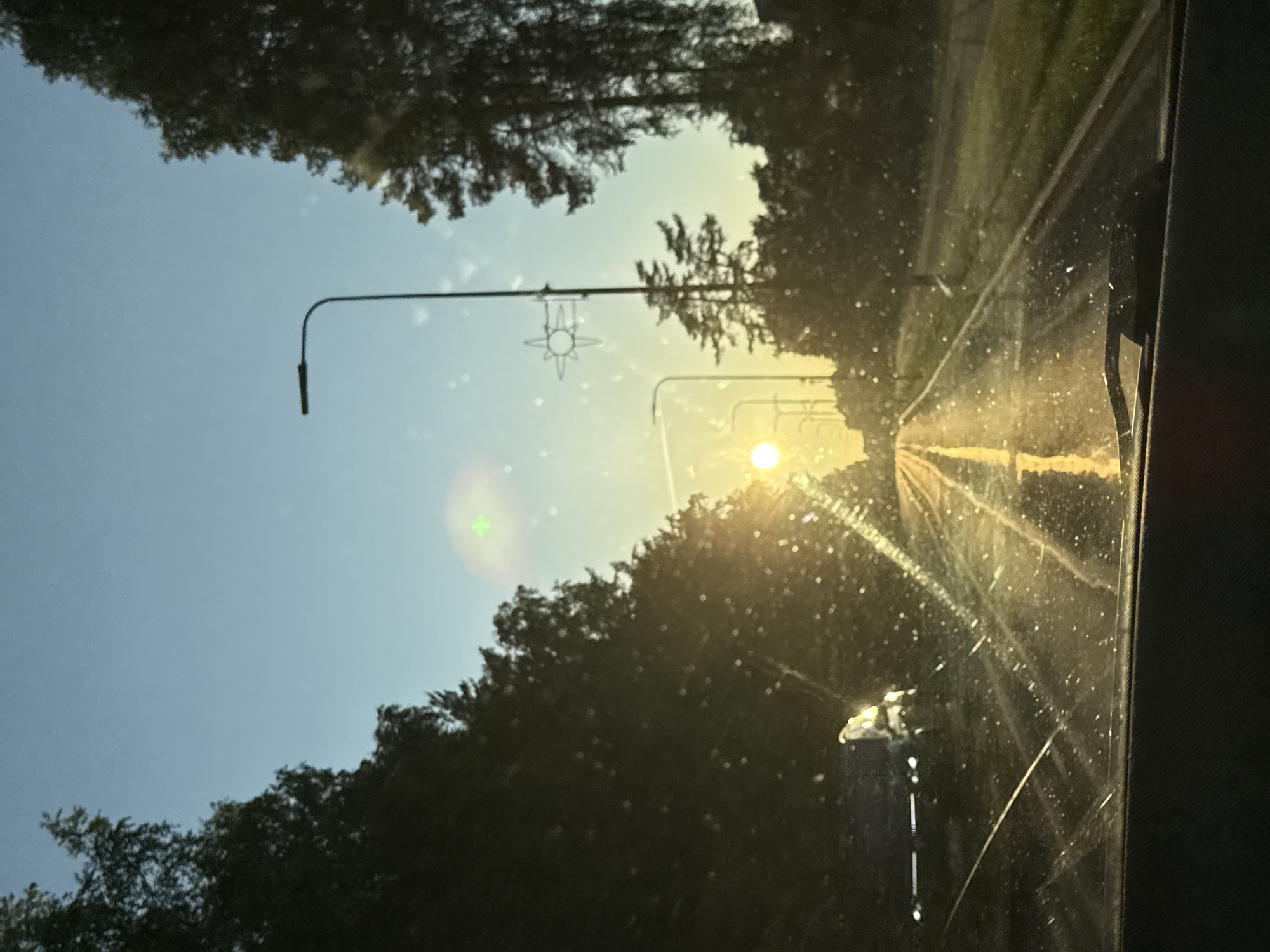 The image depicts a straight asphalt road receding into the distance, viewed from inside a vehicle through a partially obscured windshield. The windshield is covered in numerous small particles or droplets, causing light dispersion and visible streaks. A vehicle dashboard and a wiper blade are visible at the bottom. The sun is low on the horizon, positioned centrally near the vanishing point of the road, casting strong yellow light and reflections across the wet or reflective road surface, particularly along the prominent yellow central line. The sky exhibits a gradient from light blue overhead to bright yellow and orange near the sun. Dense, dark green trees line both sides of the road, silhouetted against the bright sky. Multiple streetlights with dark, curved poles are visible along the road. A distinctive star-shaped decorative element is attached to a streetlight pole on the right side of the road. Faint light sources, potentially headlights or reflections from another vehicle, are visible on the left side of the road in the mid-ground. The scene is located in the city of Tallinn, Estonia, during either sunrise or sunset. No individuals are discernible within the frame.