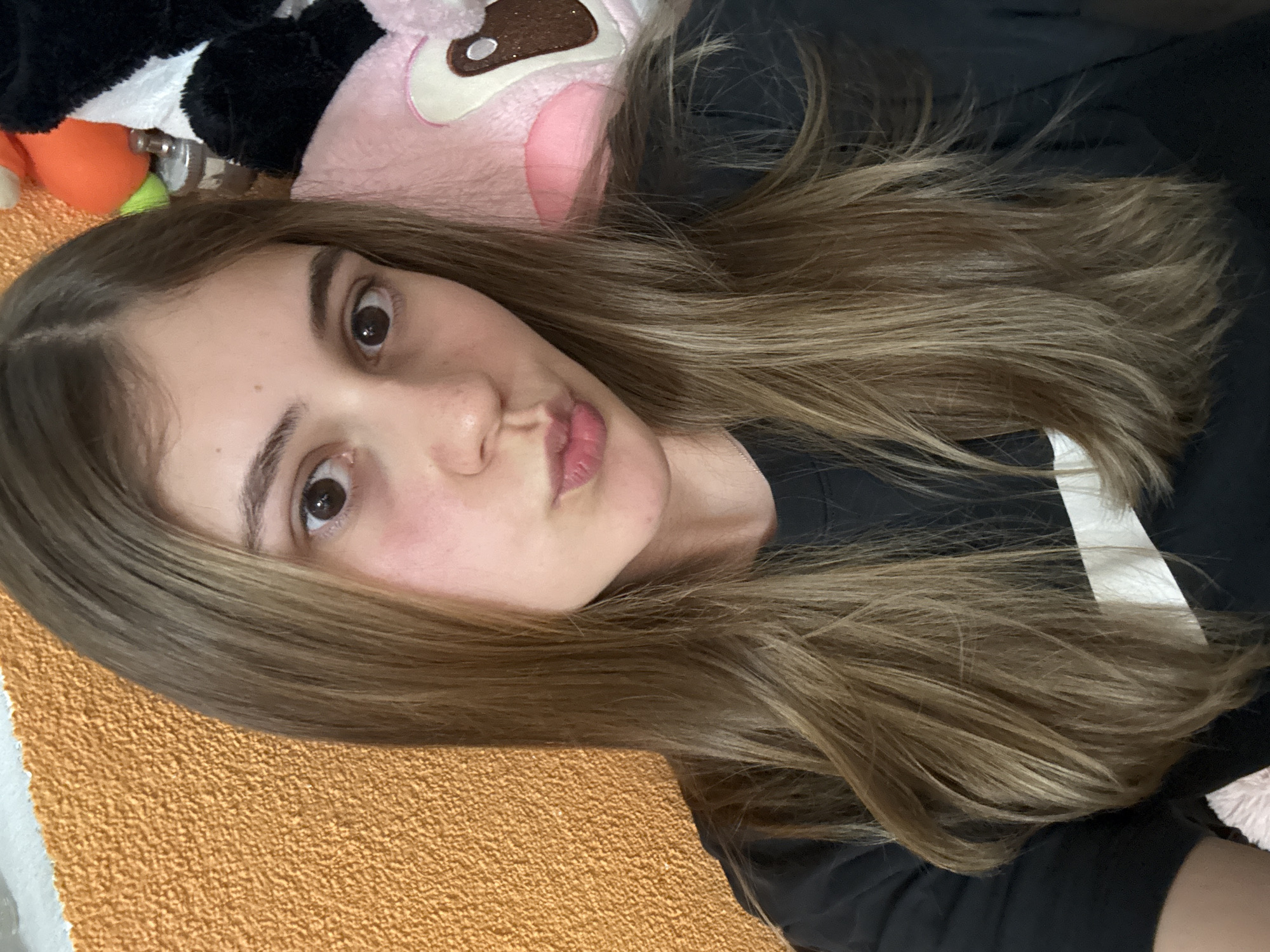 A close-up selfie of a young woman with long, light brown hair. She is looking directly at the camera with slightly puckered lips. Her eyes are brown. She is wearing a black t-shirt with a white stripe on the shoulder. To the left of her head are stuffed toys of different colours. The background appears to be an orange blanket or fabric. The location is unknown and cannot be determined from the image. No information suggests that it is in Panagyurishte, Bulgaria.