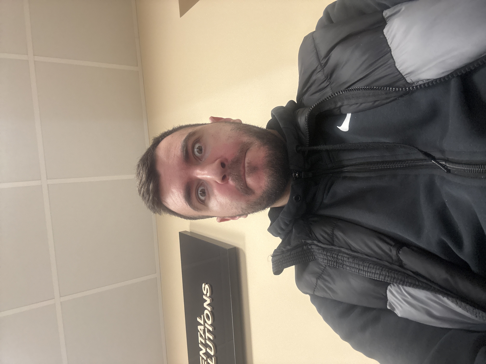 A man with short graying hair and a beard is standing inside what appears to be a dental office. He is wearing a black Nike hoodie and a black and gray puffer jacket. The background consists of a beige wall and a black sign with gold lettering that reads "DENTAL SOLUTIONS." The location is possibly in Sofia, Bulgaria, given the presence of a business operating under a common name that has dental facilities in that city.