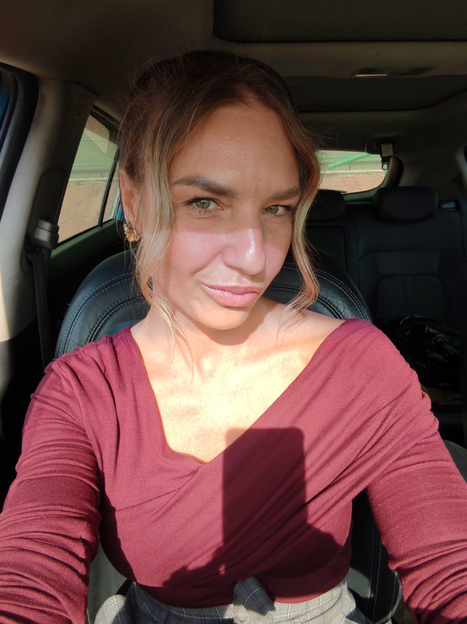 A woman with fair skin and blonde-highlighted brown hair is sitting in the driver's seat of a car. She is wearing a burgundy long-sleeved shirt. The interior of the car is visible, including black leather seats and the car's dashboard. The woman is looking directly at the camera with a slight smile. The background suggests an urban environment, but the specific location in Petropavlovsk-Kamchatsky, Russia, is not discernible from the image.