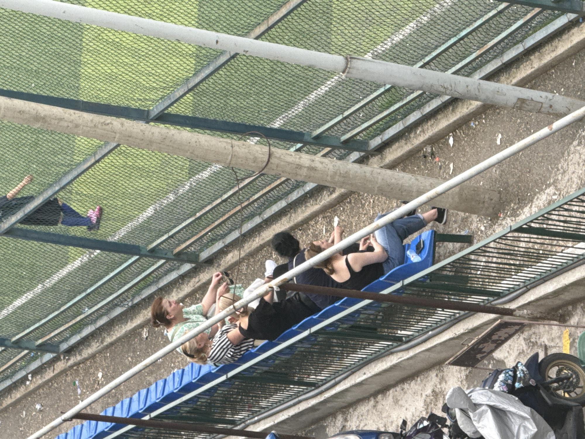 The image shows a group of people sitting on bleachers under a mesh-covered structure, possibly a sports facility. The setting appears to be outdoors, with a green field and white markings visible through the mesh, suggesting a sports field or track. The ground below the bleachers is paved with concrete and debris.
There are at least five people visible, sitting on blue and green bleachers. Some individuals are holding phones or other small objects. One person is wearing pink shoes. The overall mood seems relaxed, with people in casual attire. The lighting suggests it is daytime, but the weather is unclear.
There are some visible objects in the foreground and background. In the lower right corner, parts of two motorcycles are visible, one with a black and yellow wheel and another partially covered by a silver tarp. Bags are also present near the motorcycles.
There is no visible text in the image. The scene is shot from an elevated angle, looking down on the people and the surroundings. The location context of Tbilisi, Georgia, doesn't offer specific visual cues within the image itself, but it sets a general geographic understanding.