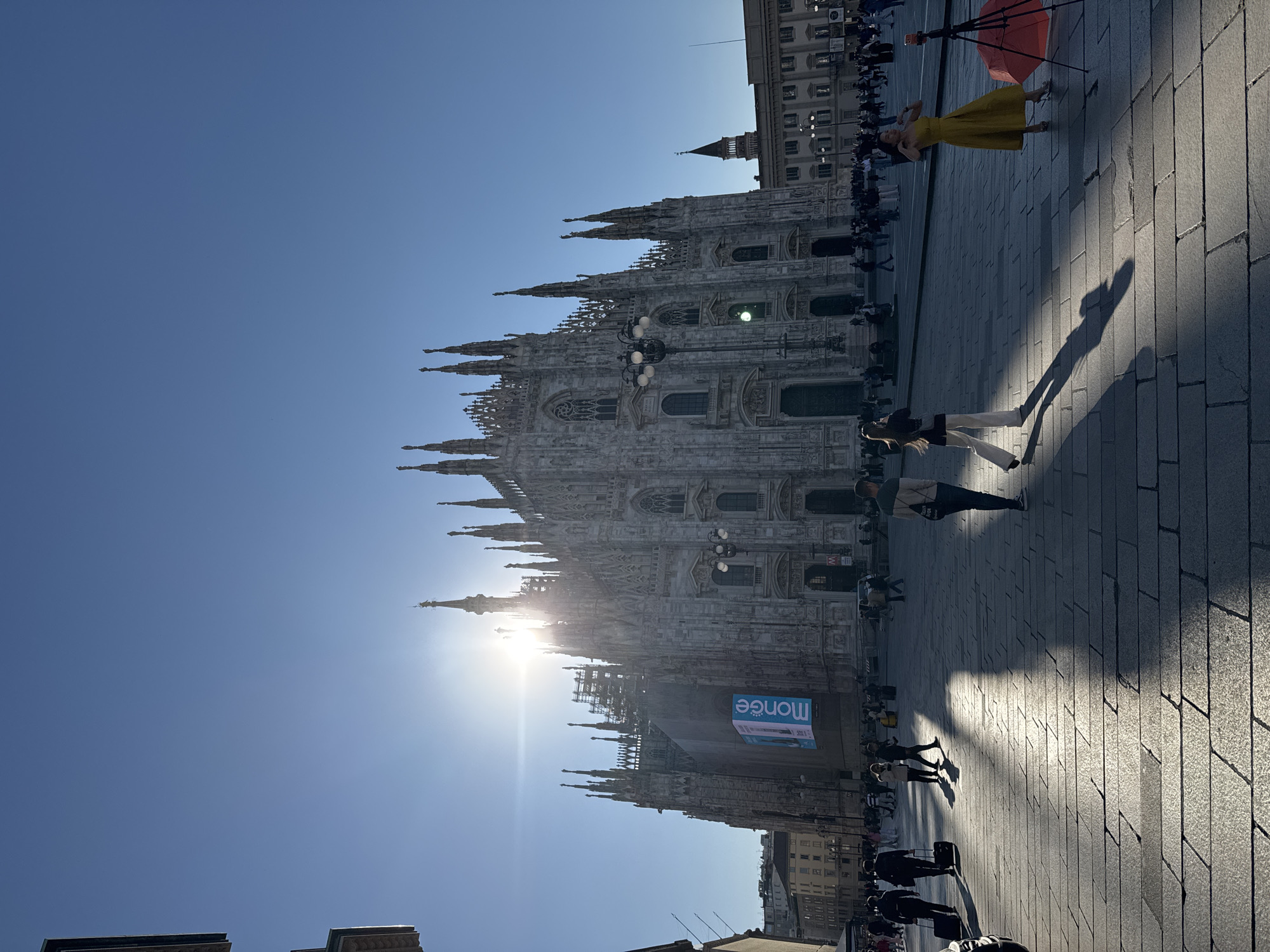 The image captures the Duomo di Milano in Milan, Italy, on a sunny day. The sky is a clear blue. Sunlight glares behind the spires of the cathedral.
The cathedral dominates the frame, its elaborate facade detailed with statues and windows. A large advertisement banner hangs on the side of the cathedral.
People are walking across the paved square in front of the Duomo. Some are tourists, others are passersby. A woman in a yellow dress poses near an open red umbrella. The shadow of a person taking the photo is visible on the ground.